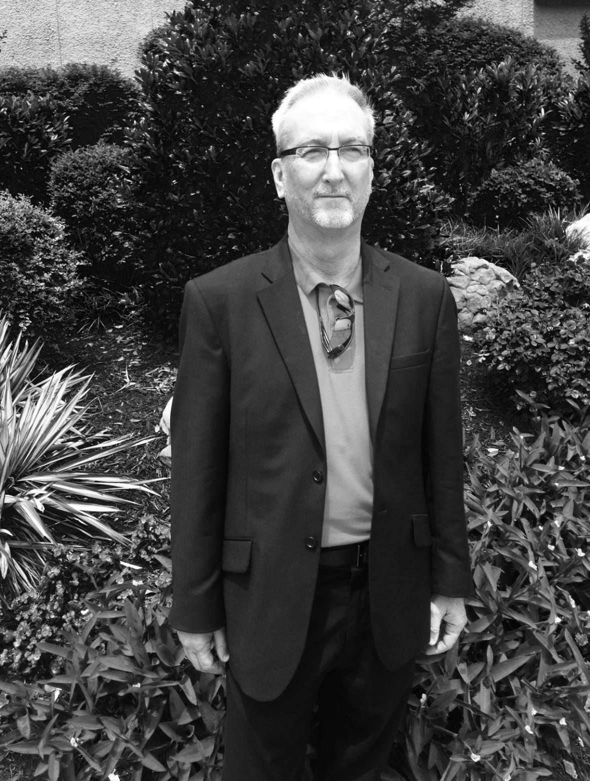 A well-dressed man poses in a serene garden surrounded by vibrant plants and trees.