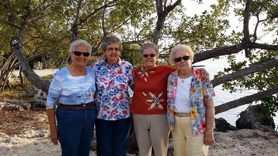 Four elderly women pose together happily against a scenic backdrop of trees and water.