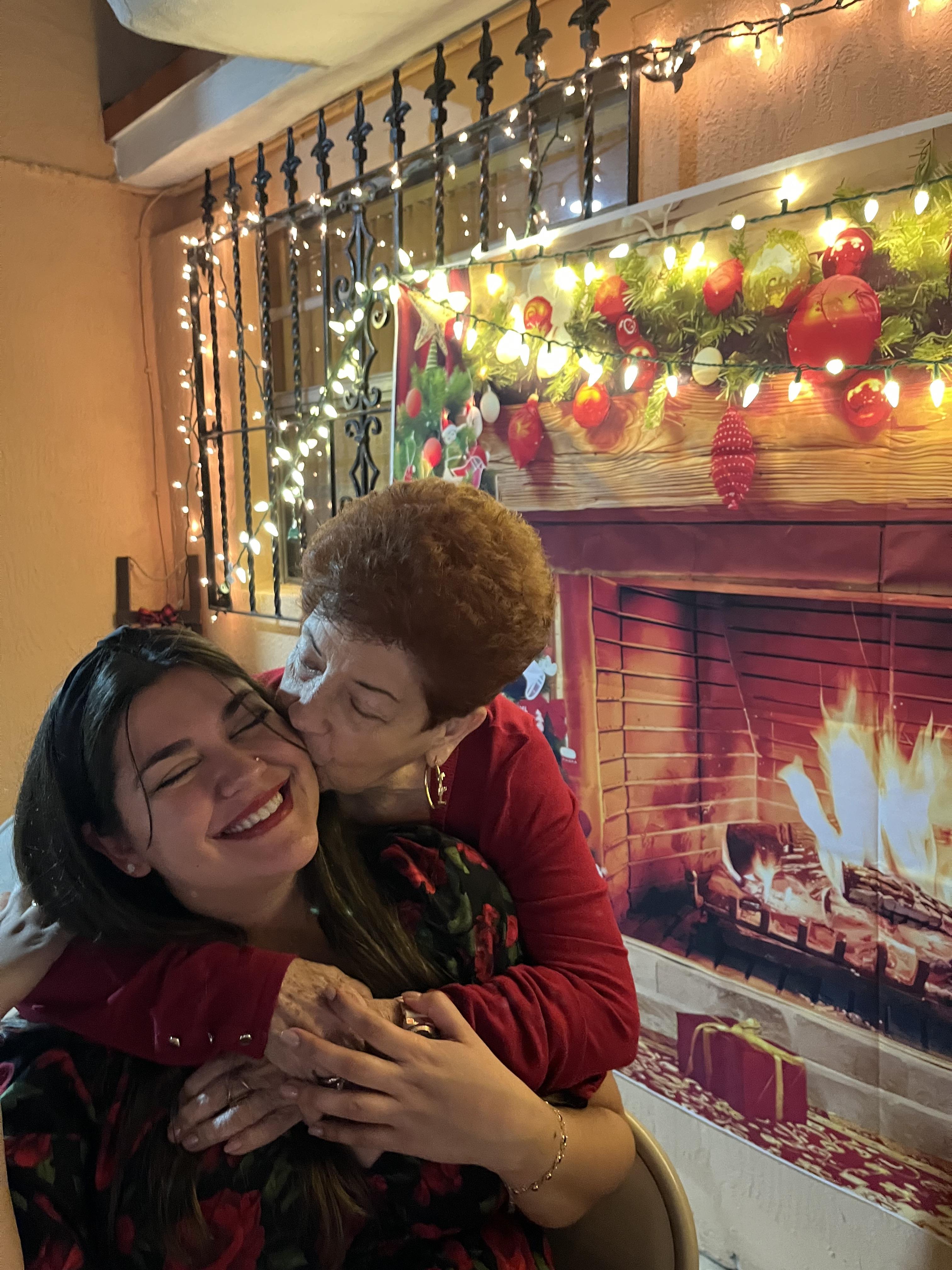 A woman kisses her smiling daughter in a warmly decorated space with festive lights.