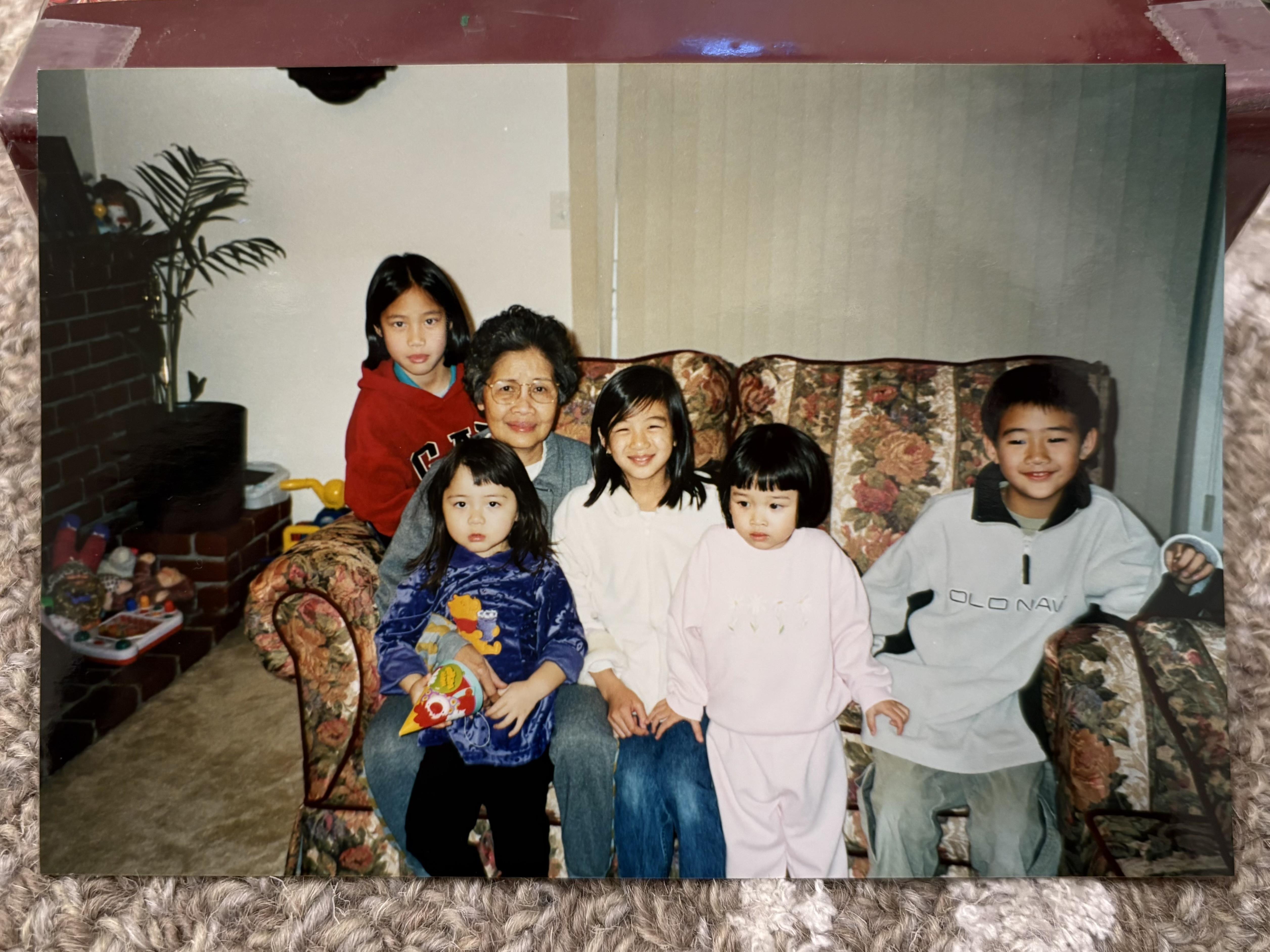 Six children sit cheerfully with their grandmother on a floral couch in a cozy living room.