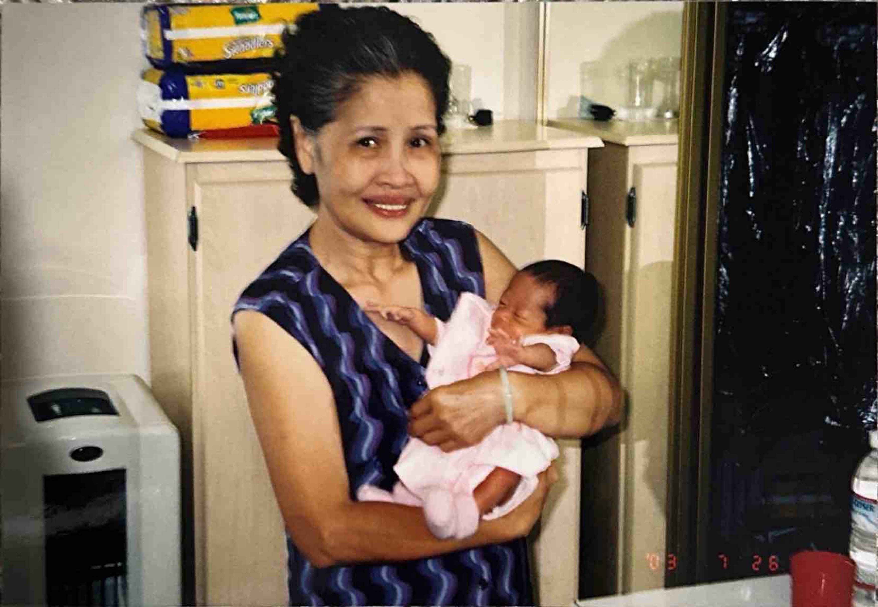 An elderly woman smiles joyfully while cradling a newborn baby in a bright indoor environment.