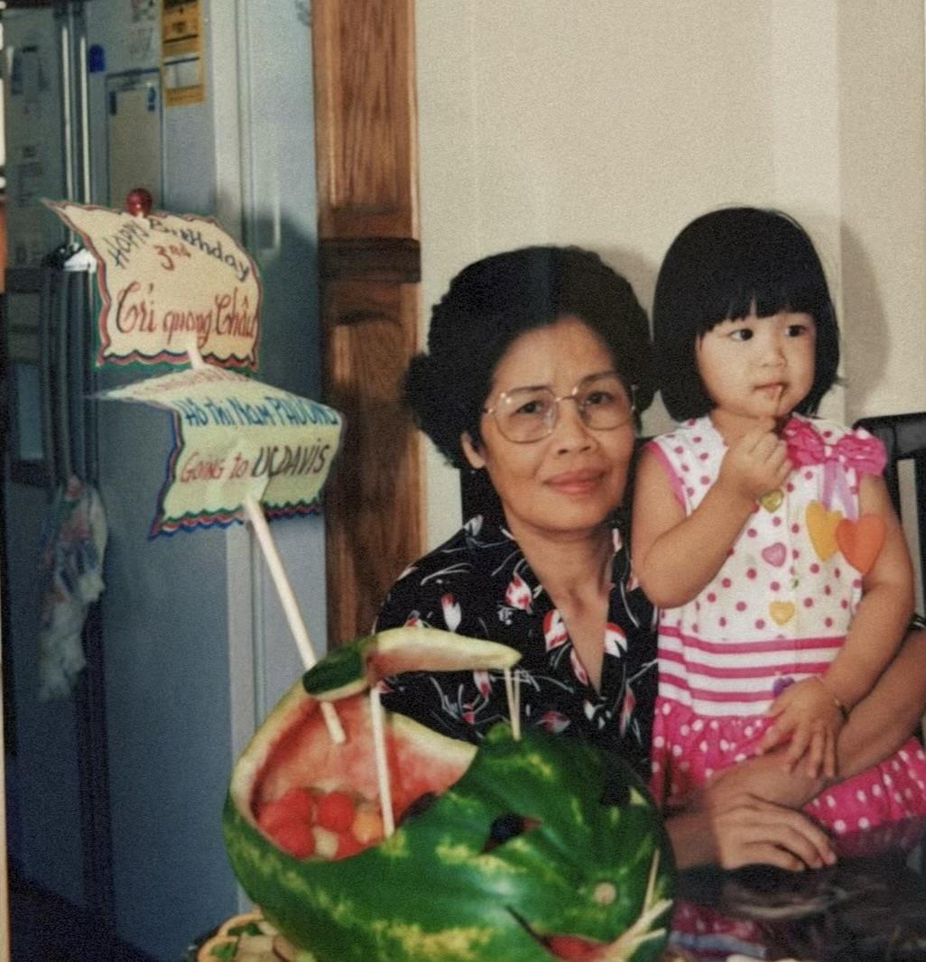 An elderly woman smiles, holding a young girl in a vibrant kitchen filled with colorful fruits.