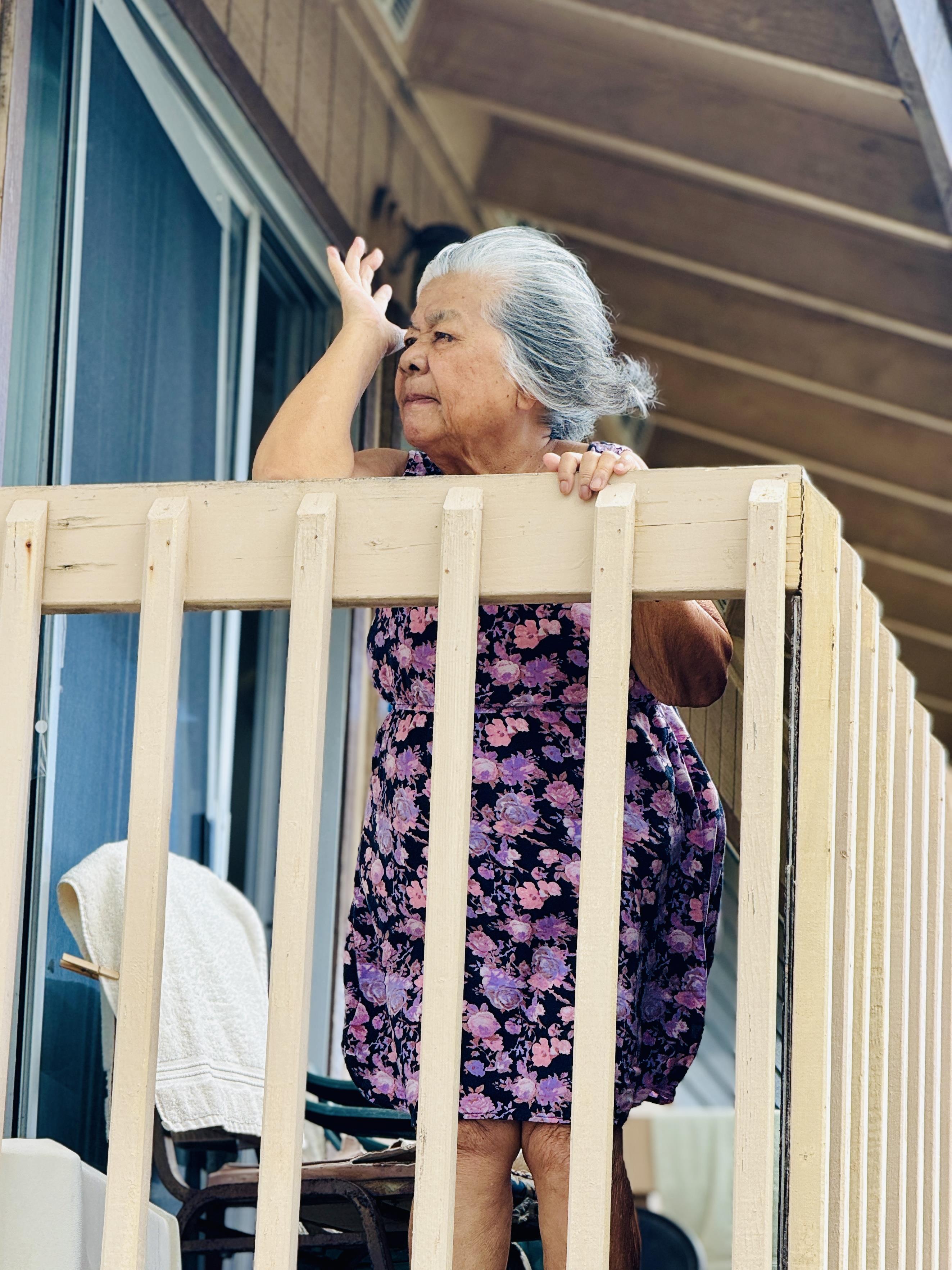 An elderly woman is peering out from her balcony, enjoying the sunshine and fresh air.