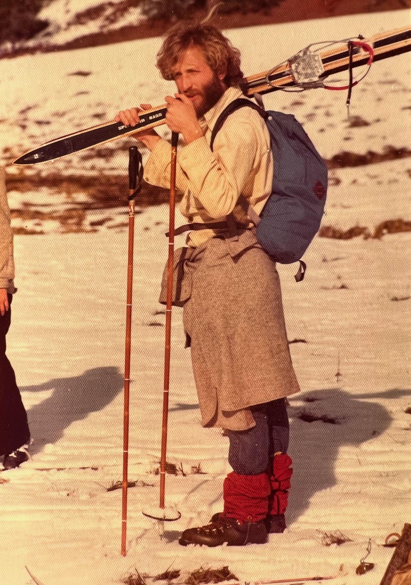 A skier stands on snow-covered ground, holding skis and poles, preparing for a day in the mountains.