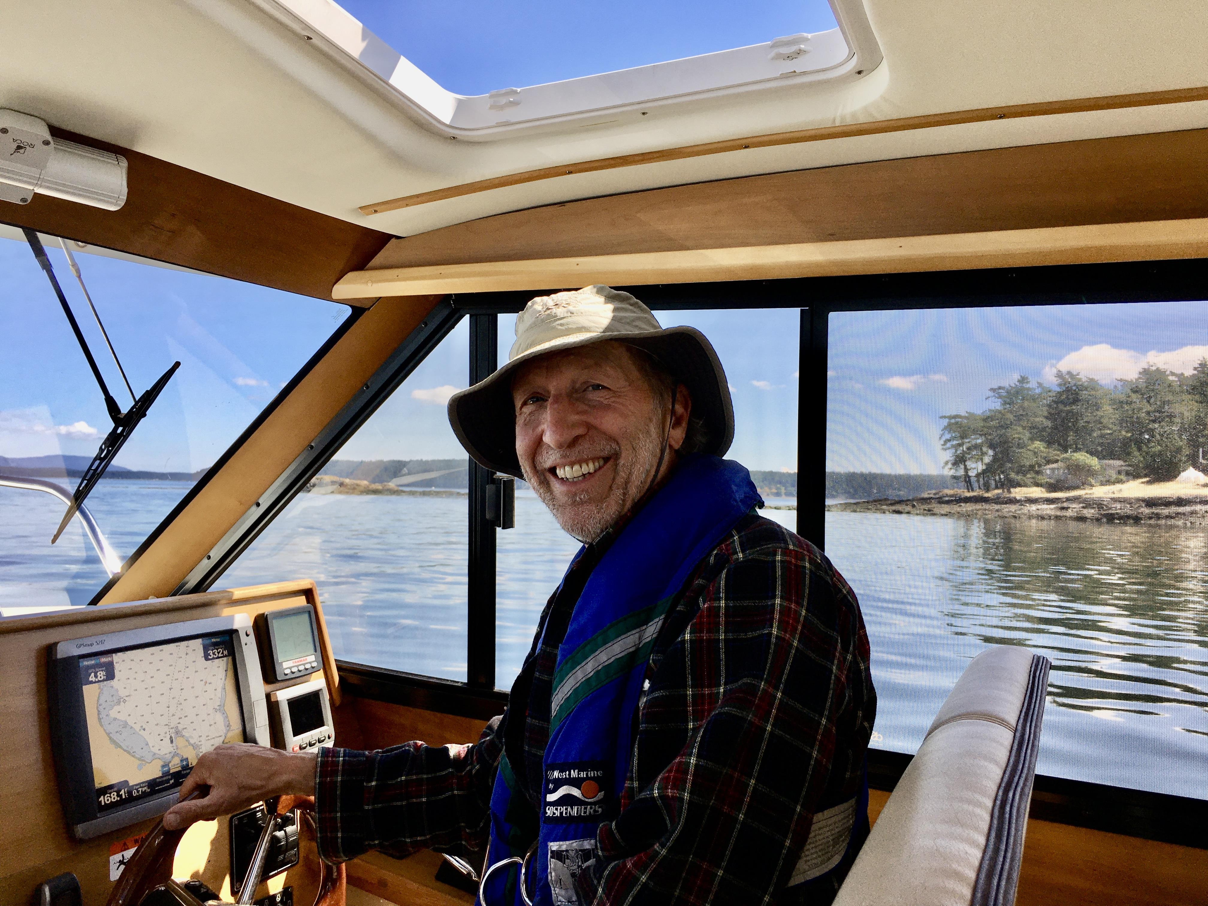 Man in a hat steers a boat on clear waters, smiling at the picturesque view under a bright sky.