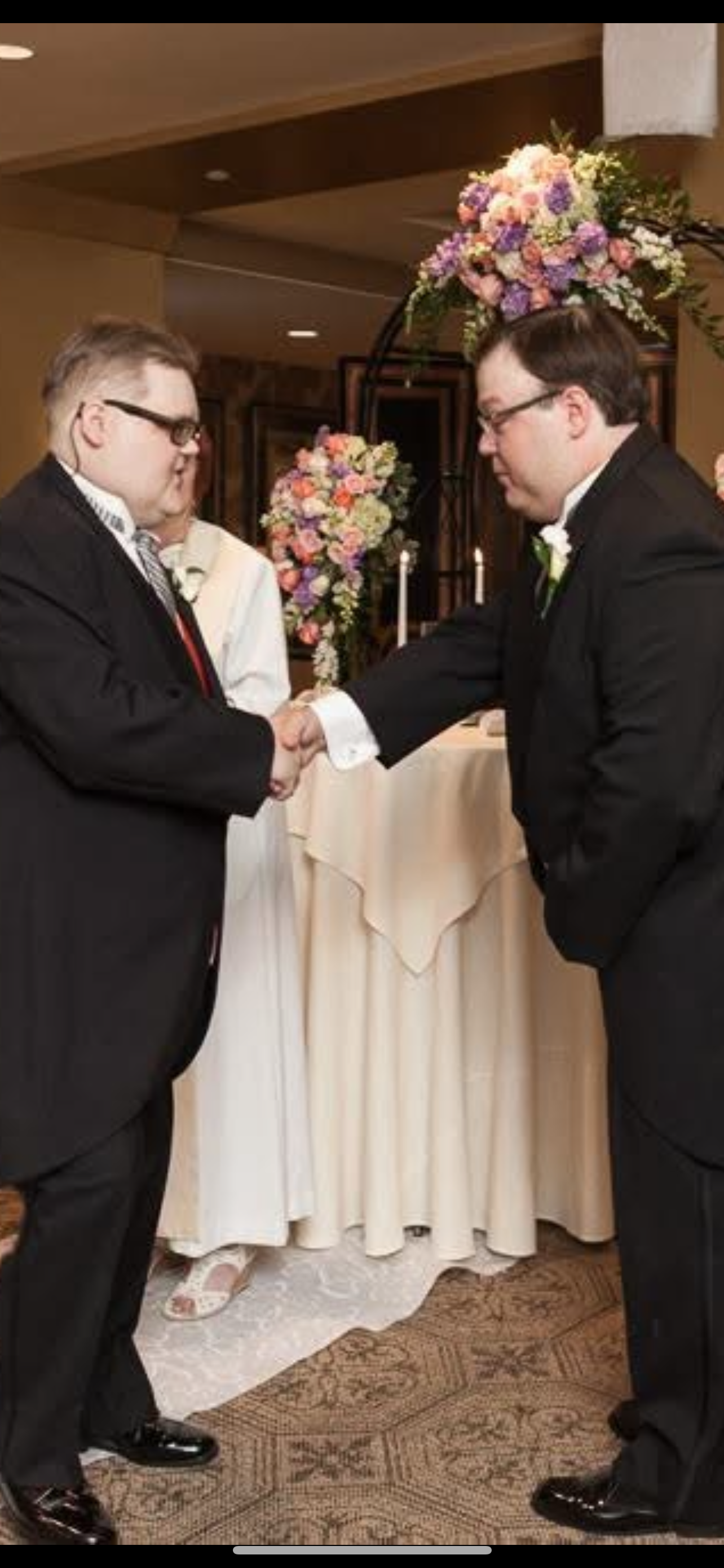Two grooms shake hands at their wedding, surrounded by guests and beautiful floral arrangements.