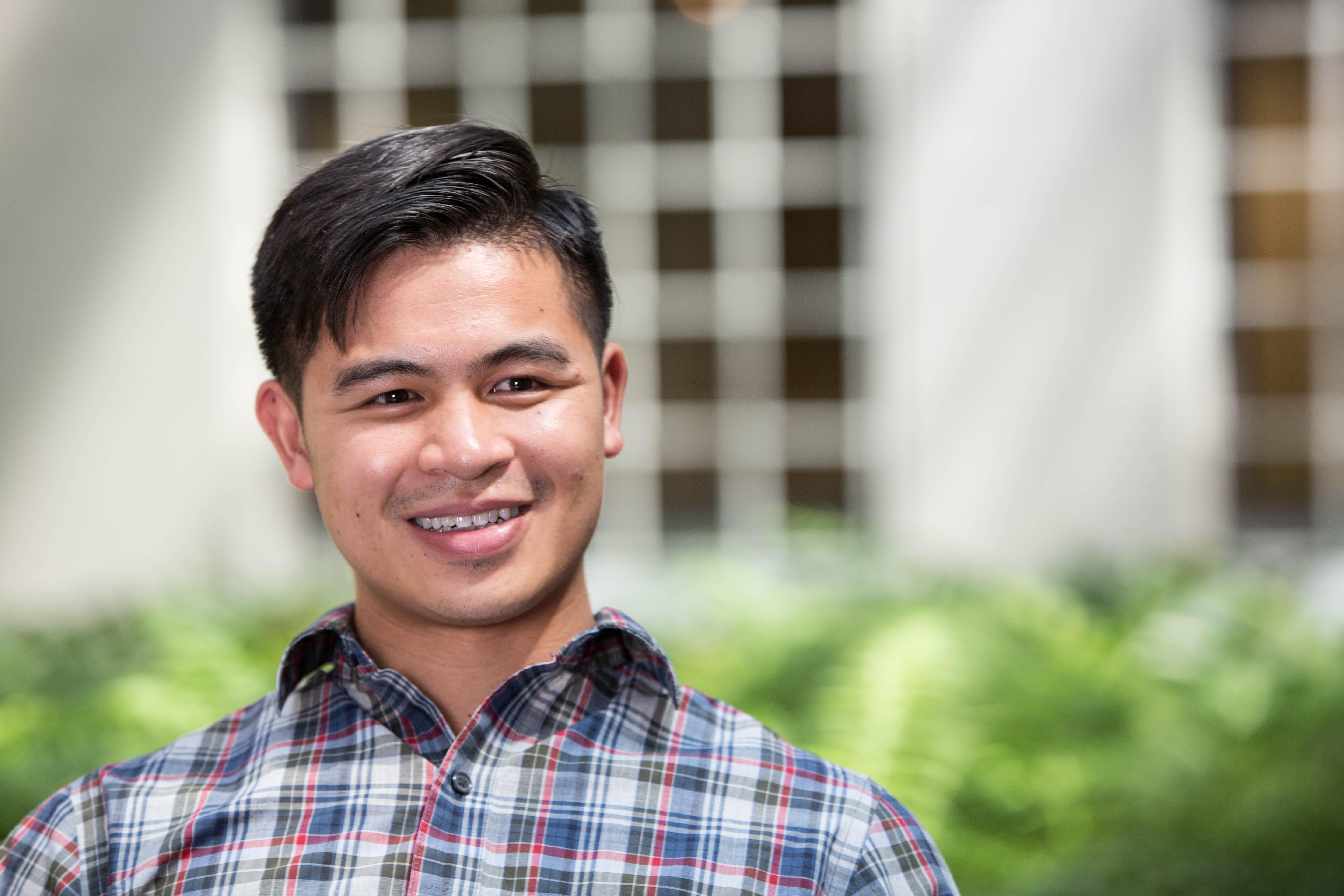 Man stands outside, smiling and dressed in a plaid shirt with greenery and a building behind him.