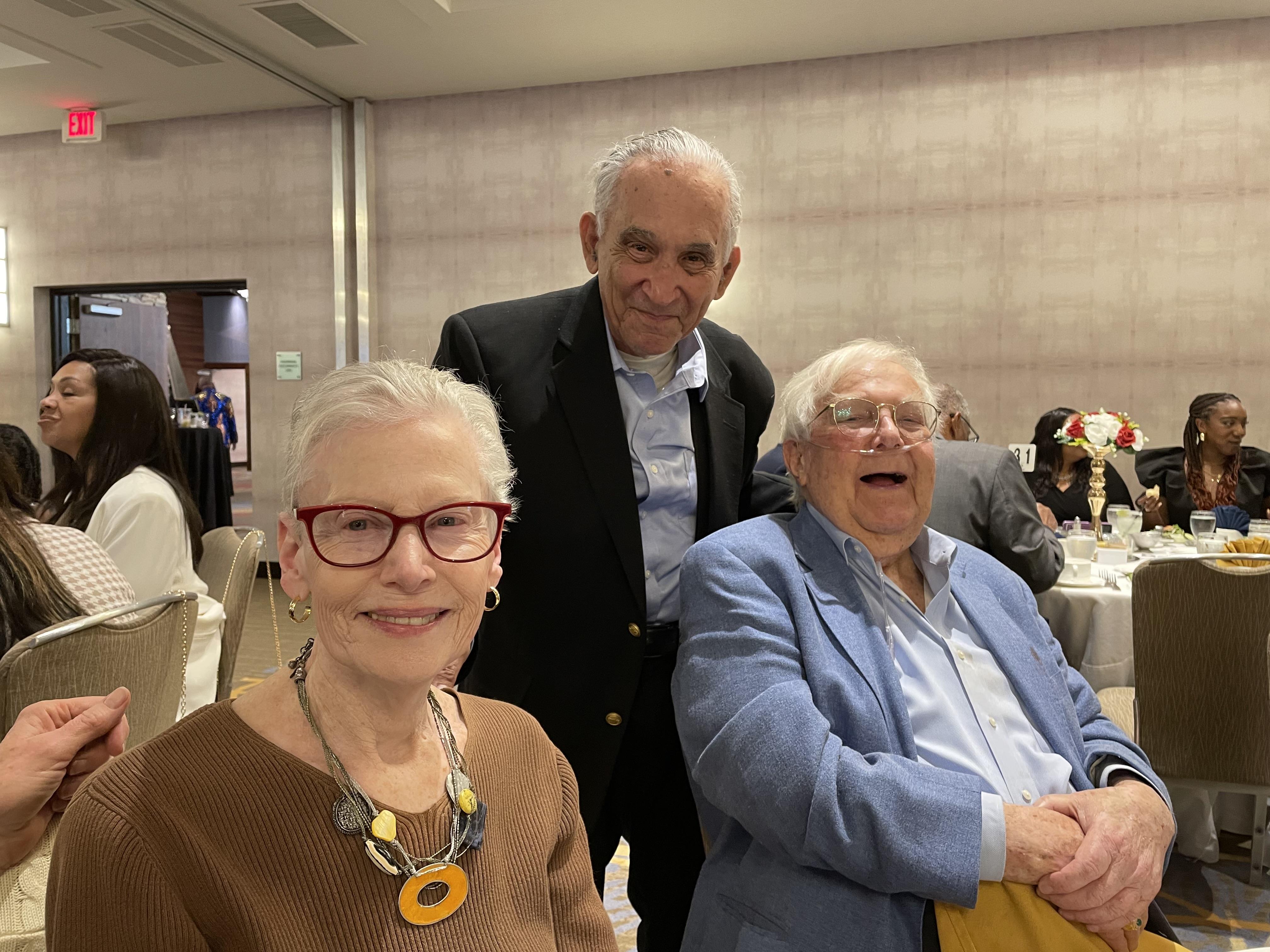 Three seniors are smiling and socializing together at a festive banquet event in a bright venue.