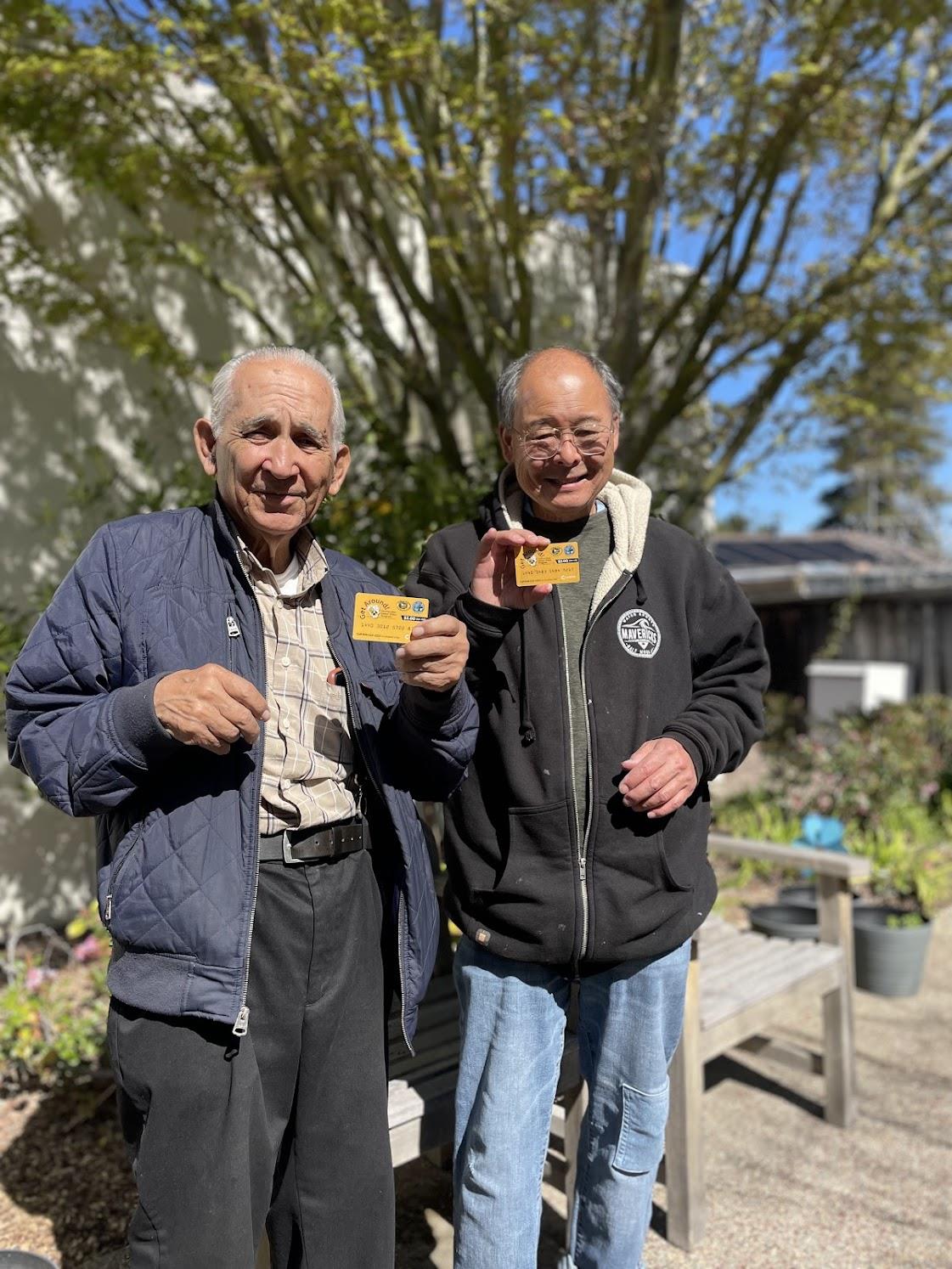 Two joyful senior men stand in a garden, proudly holding their cards while smiling at the camera.