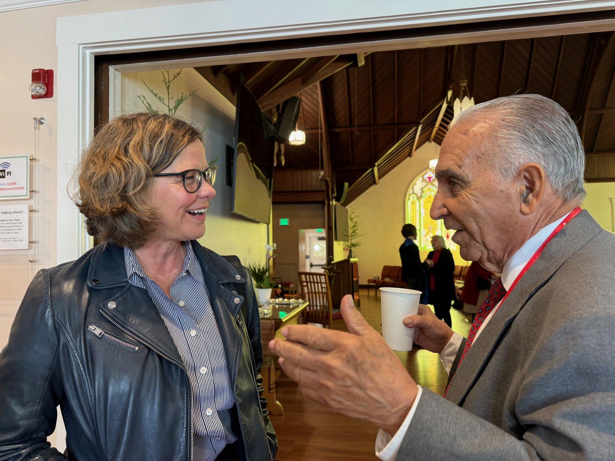 Two people chat animatedly indoors, sharing smiles and holding drinks in a welcoming environment.