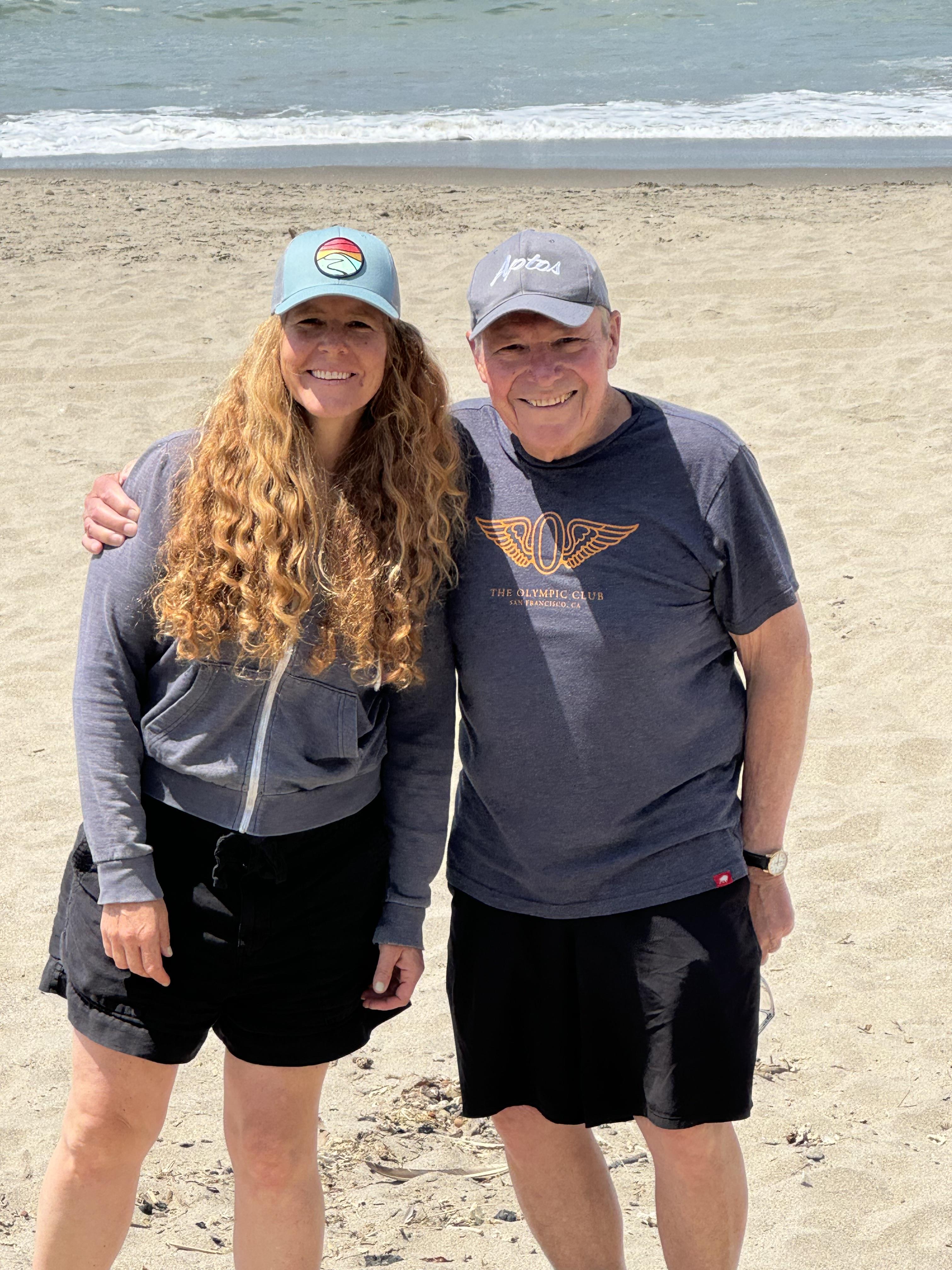 A man and woman stand together on the sand, both wearing casual clothes and hats, smiling.