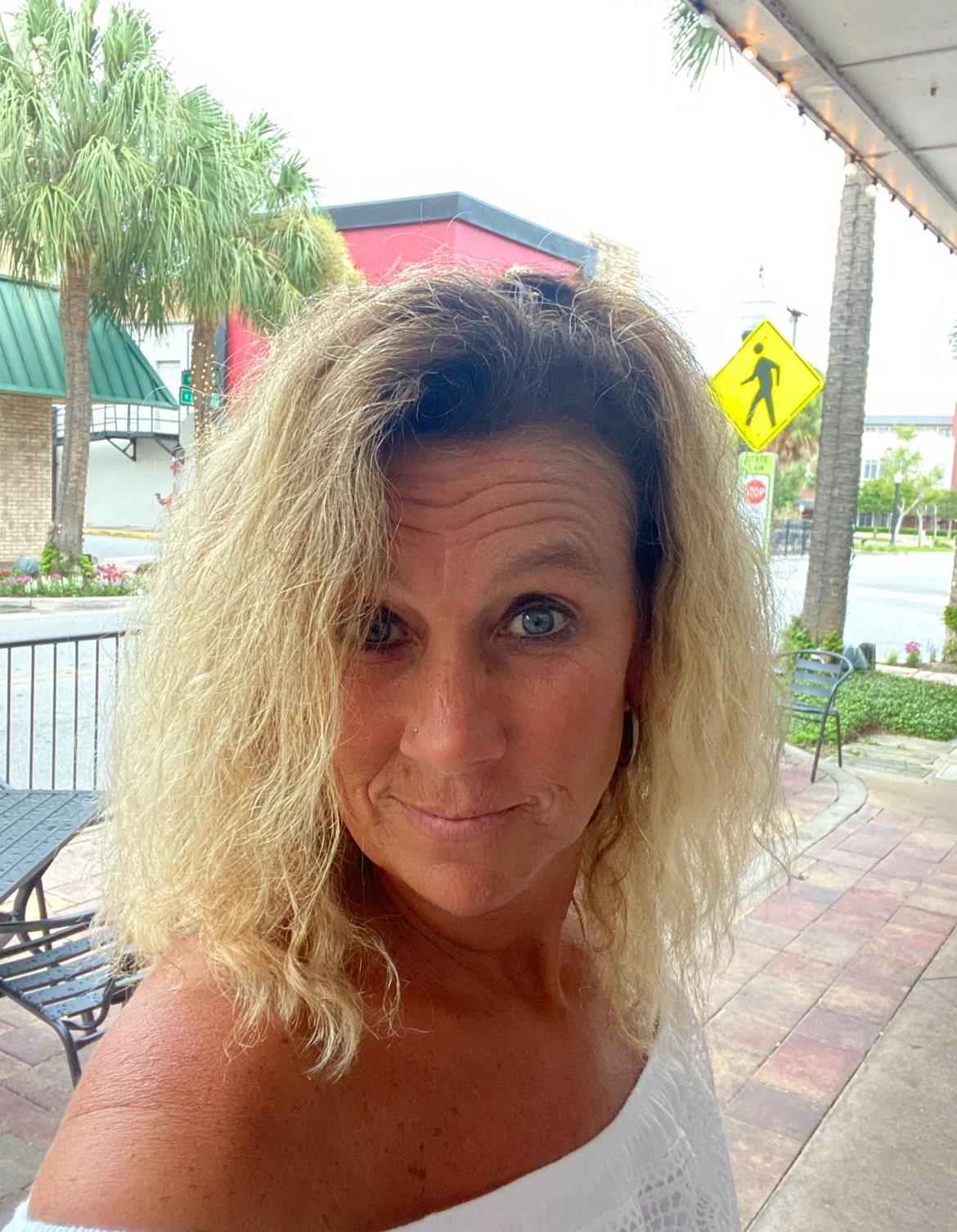 Woman with curly hair poses outdoors, enjoying a sunny day near palm trees and urban architecture.