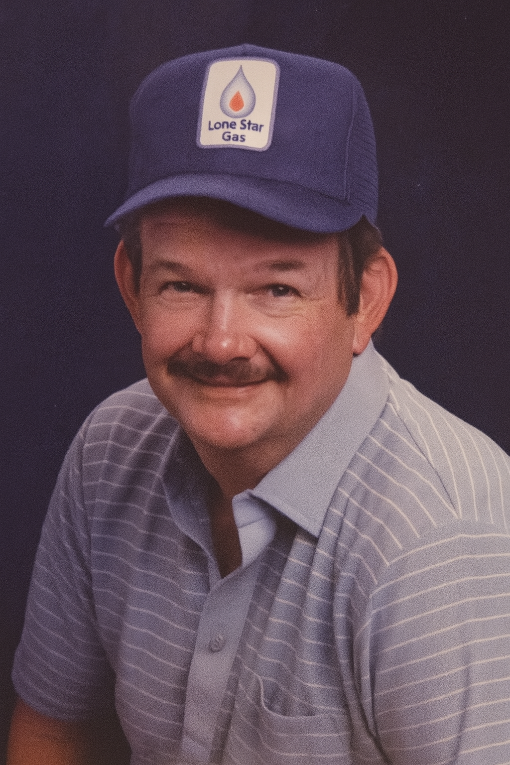 A cheerful older man in a blue cap and striped shirt poses for a portrait in a studio.