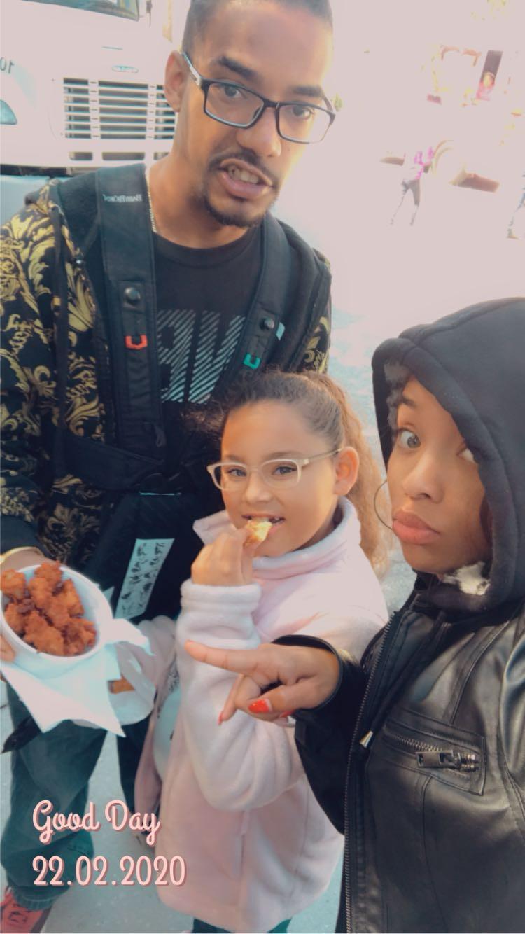 A girl tastes tasty treats as two adults pose happily during an outdoor gathering.