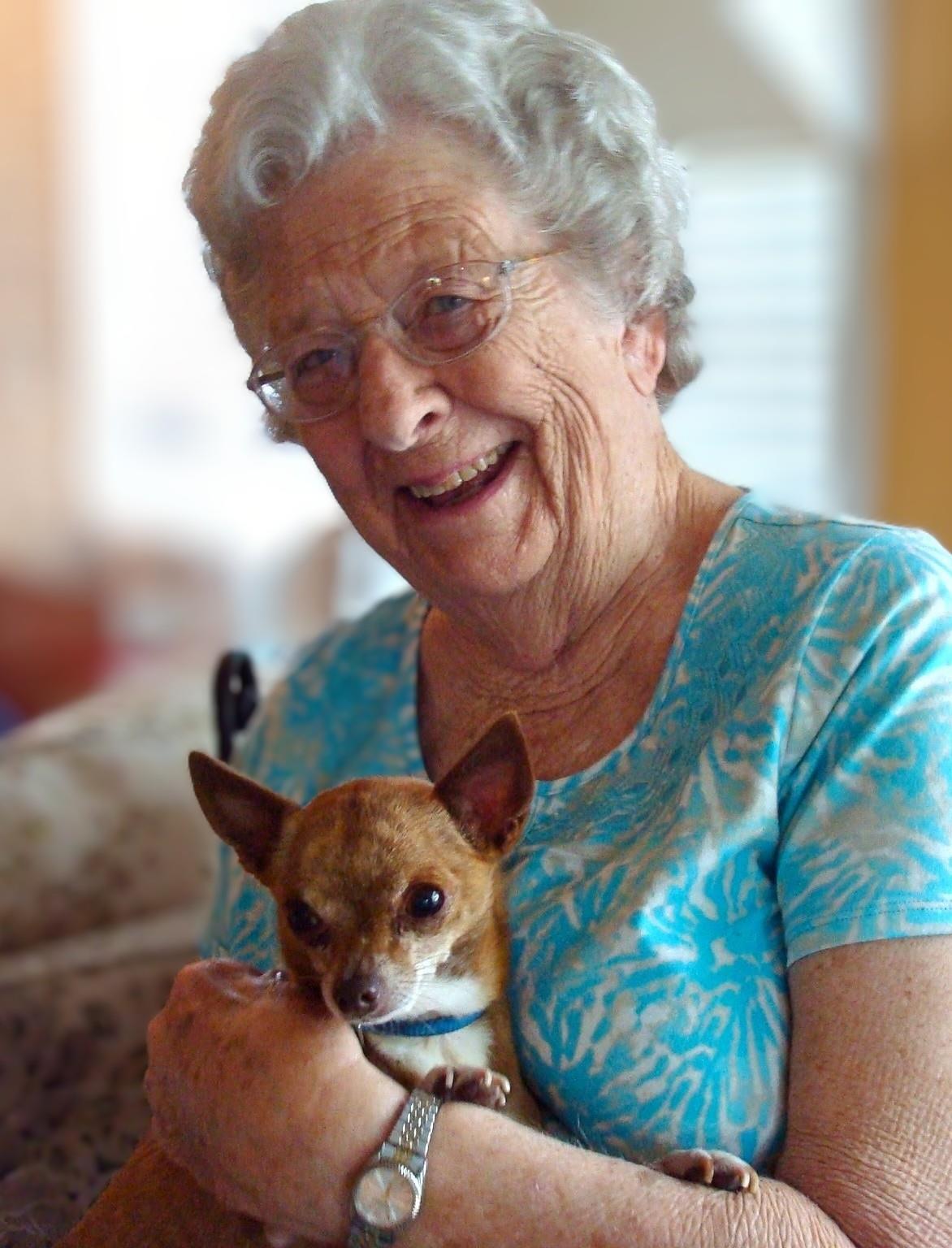 An elderly woman beams with joy as she cuddles a small dog in a warm, inviting room.