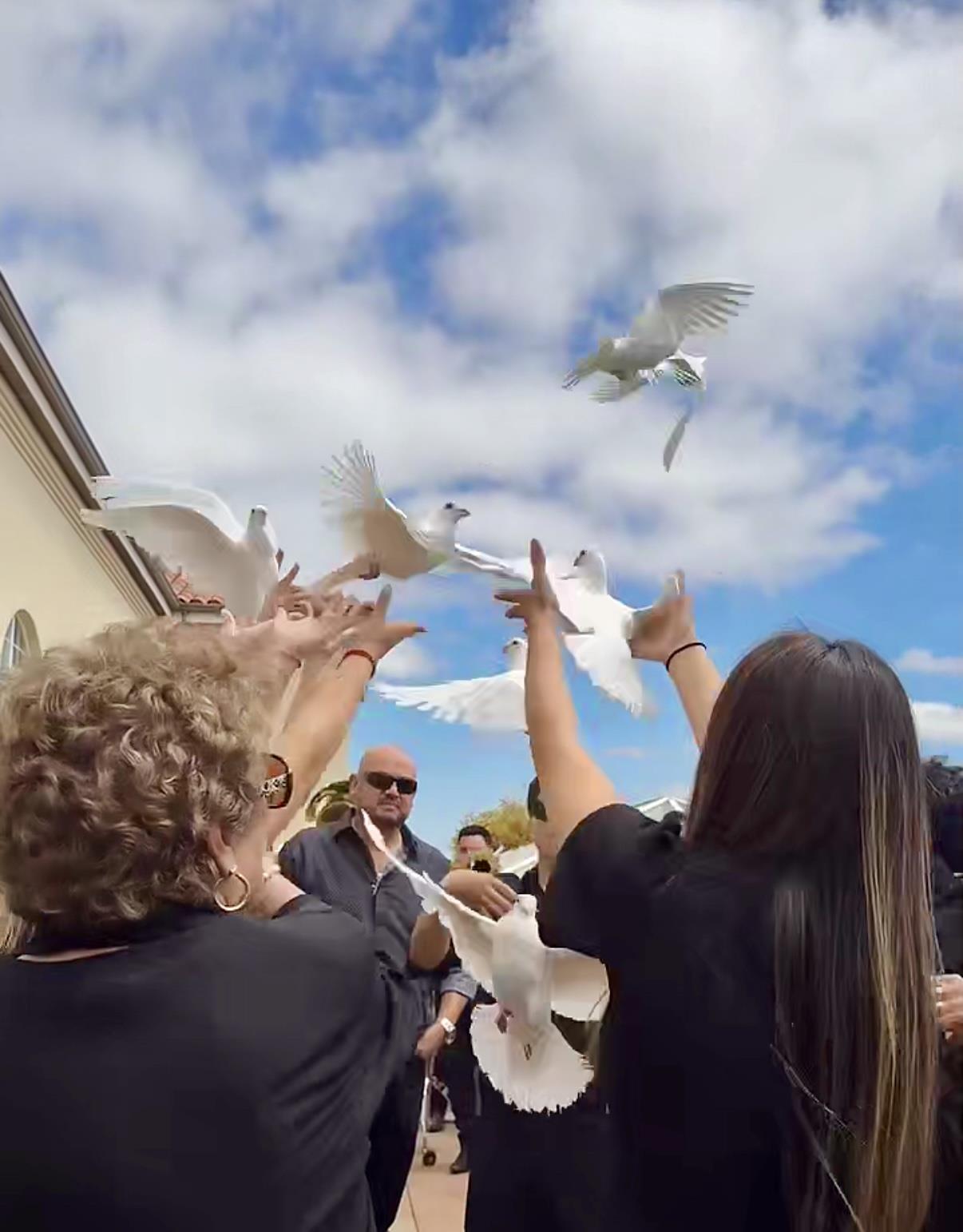 Attendees at a memorial ceremony release white doves into a clear blue sky, symbolizing peace.