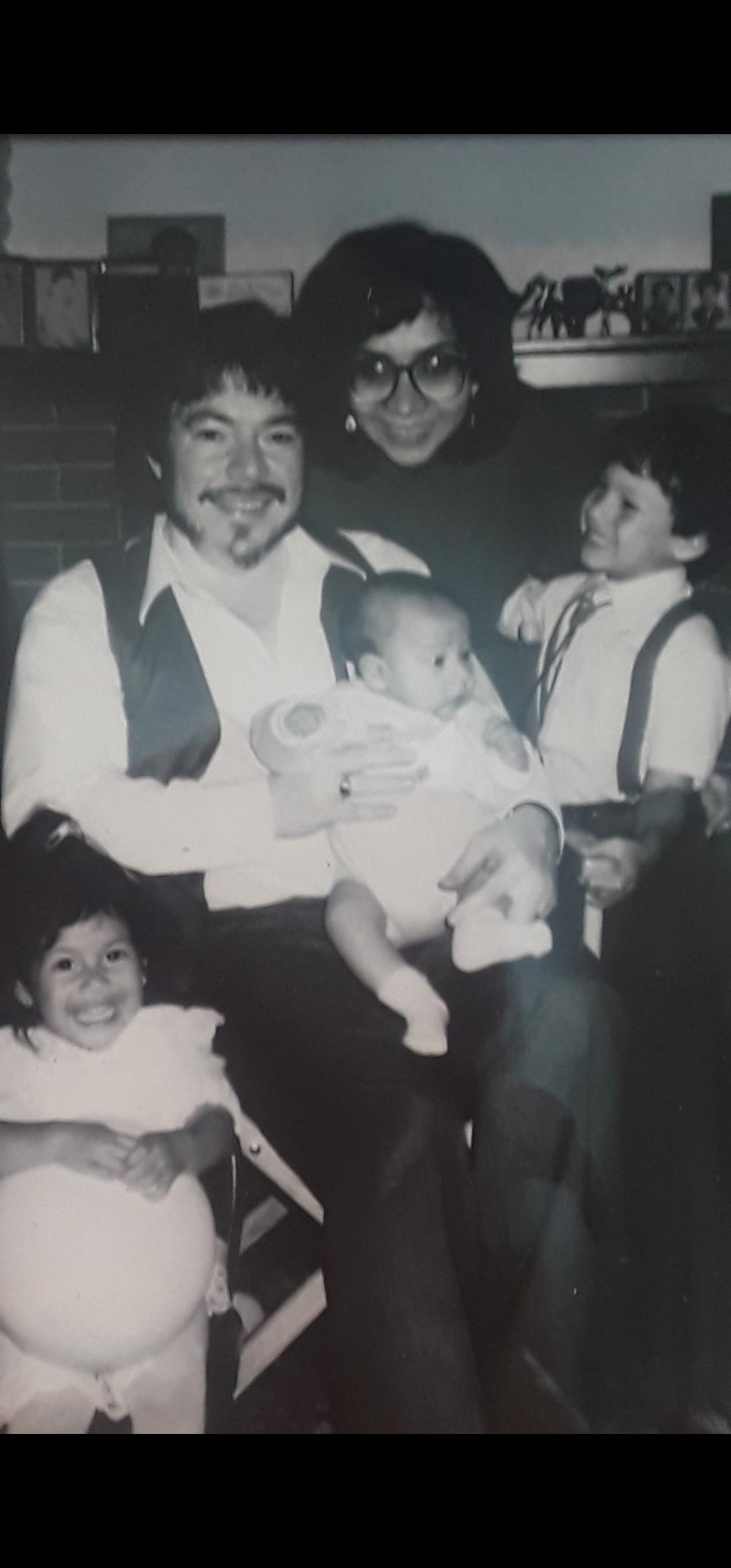 A cheerful adult holds a baby while two children sit nearby, smiling happily in a warm setting.