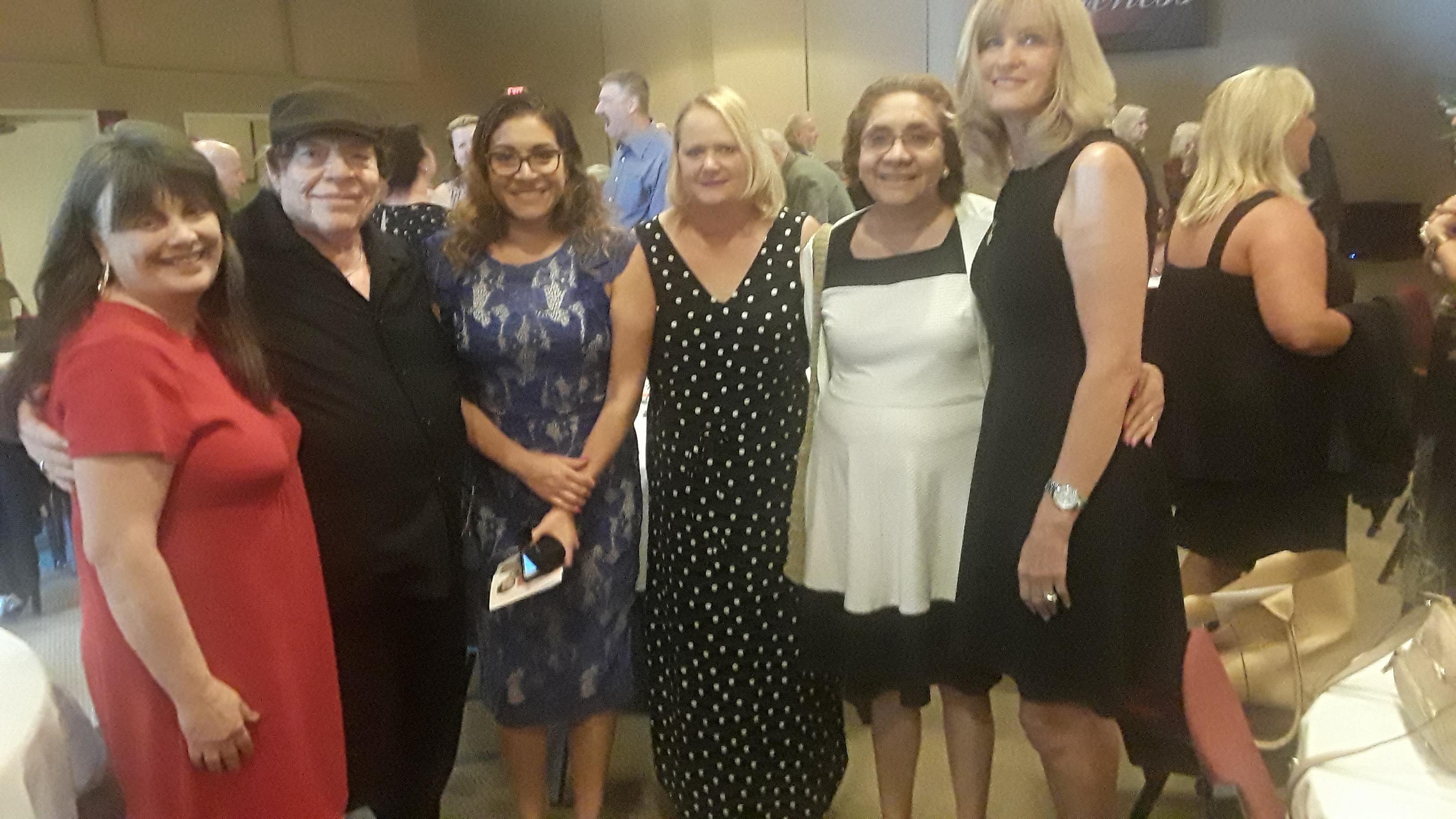 A group of five women stand together, smiling and enjoying their time at a social gathering.