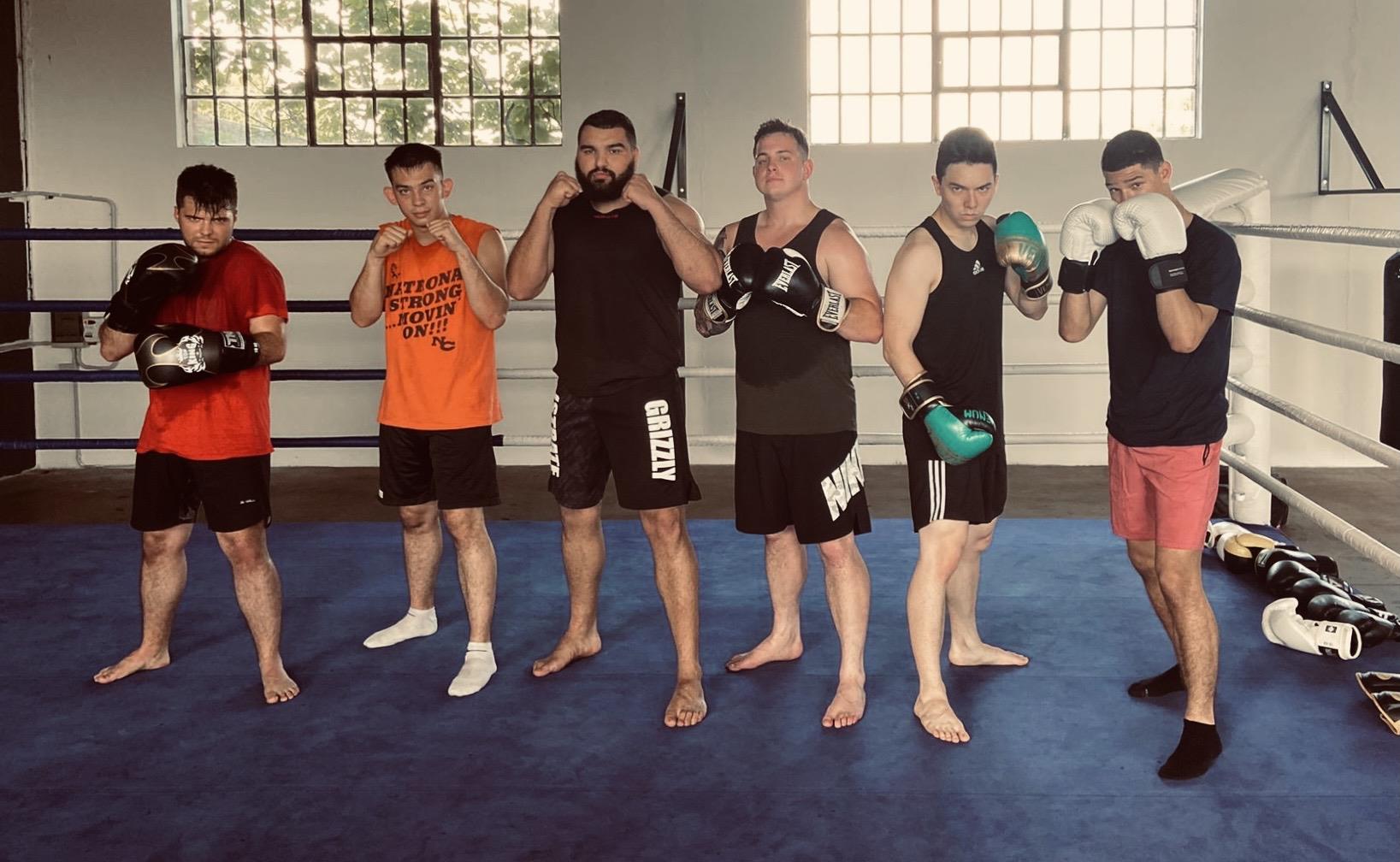 Six martial artists with boxing gloves pose in a gym, showcasing focus and camaraderie.