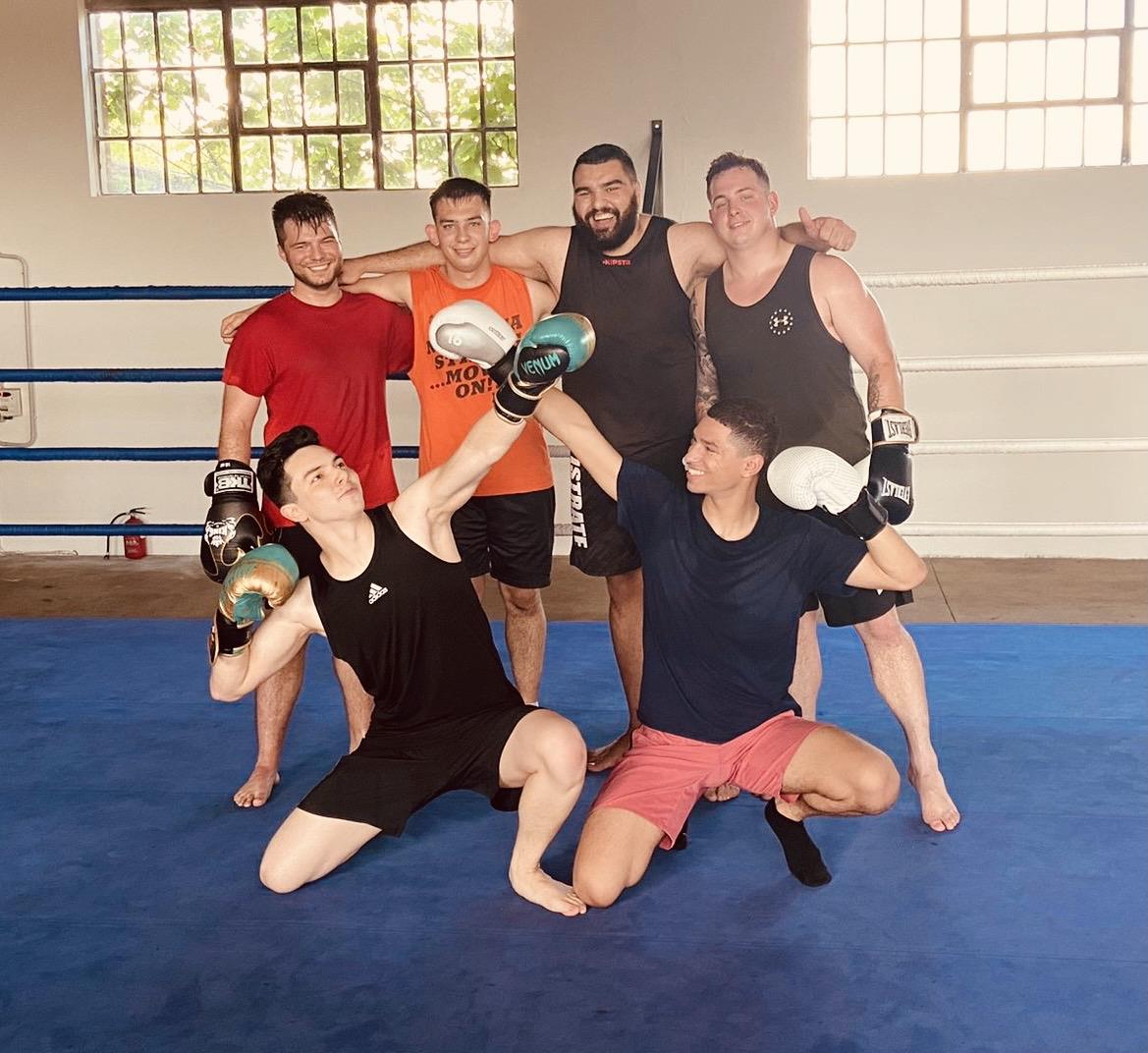 Six men pose together in a boxing ring, celebrating their training session with excitement.