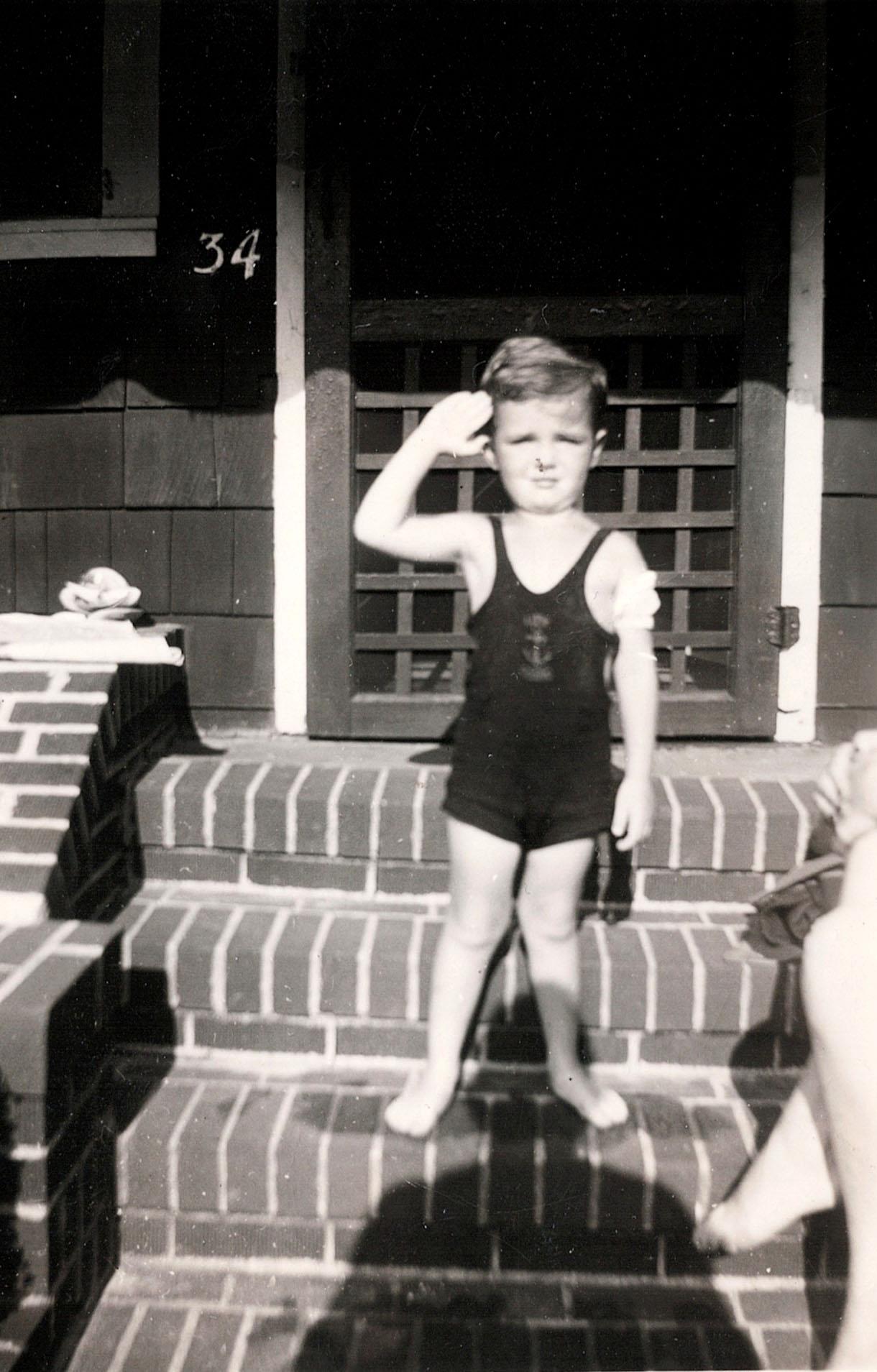 A boy wearing a swimsuit stands at the steps of a house, saluting with a serious expression.