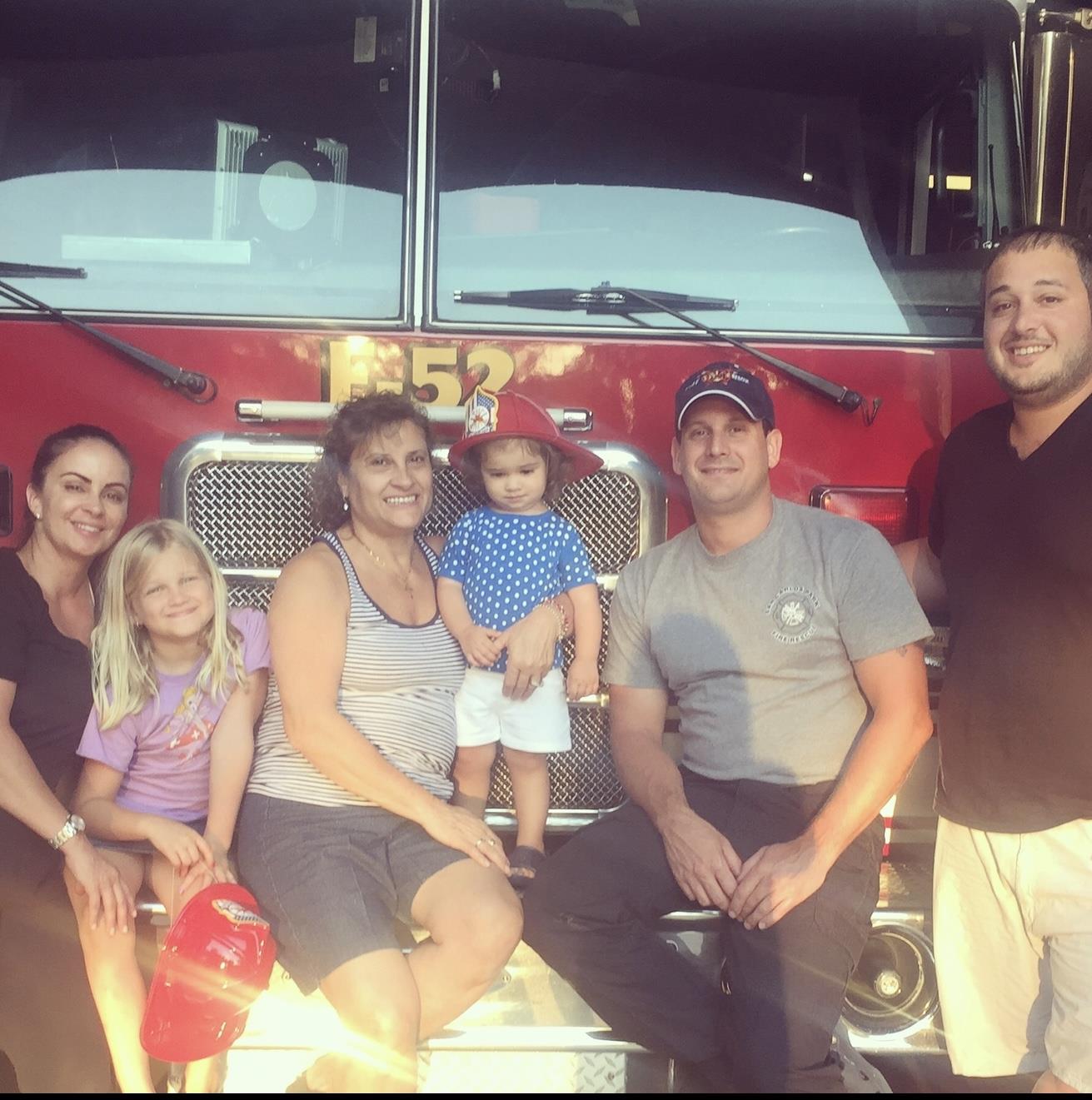 A cheerful family gathers in front of a fire truck during a summer evening community event.