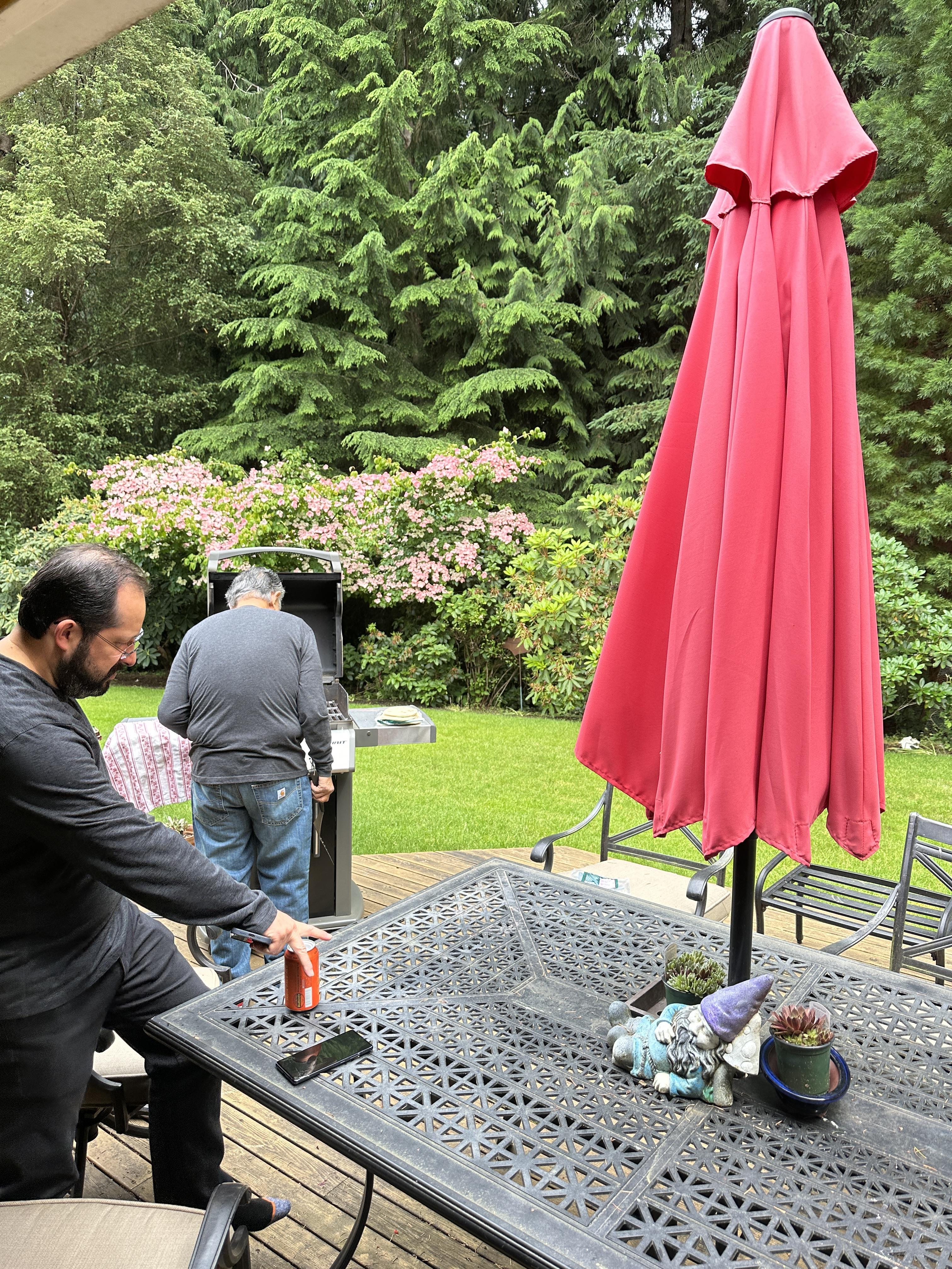 Two men enjoy a casual meeting outdoors, preparing food at a grill surrounded by greenery.