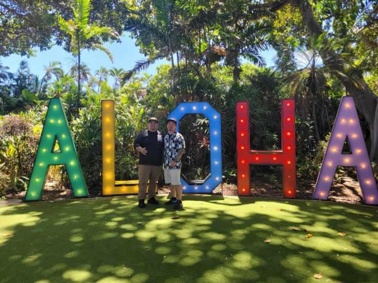Two friends stand by a large illuminated Aloha sign surrounded by lush greenery in Hawaii.