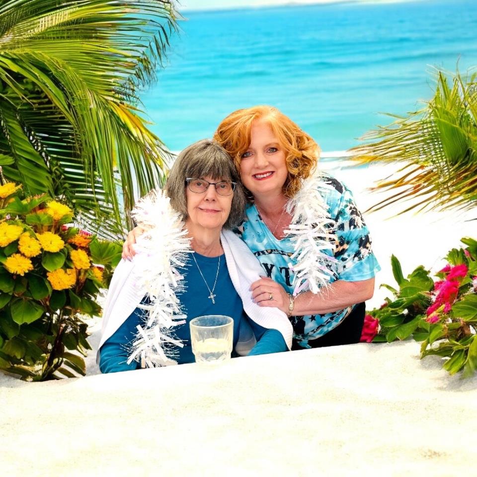 Two women smile and embrace in a tropical paradise filled with vibrant flora.