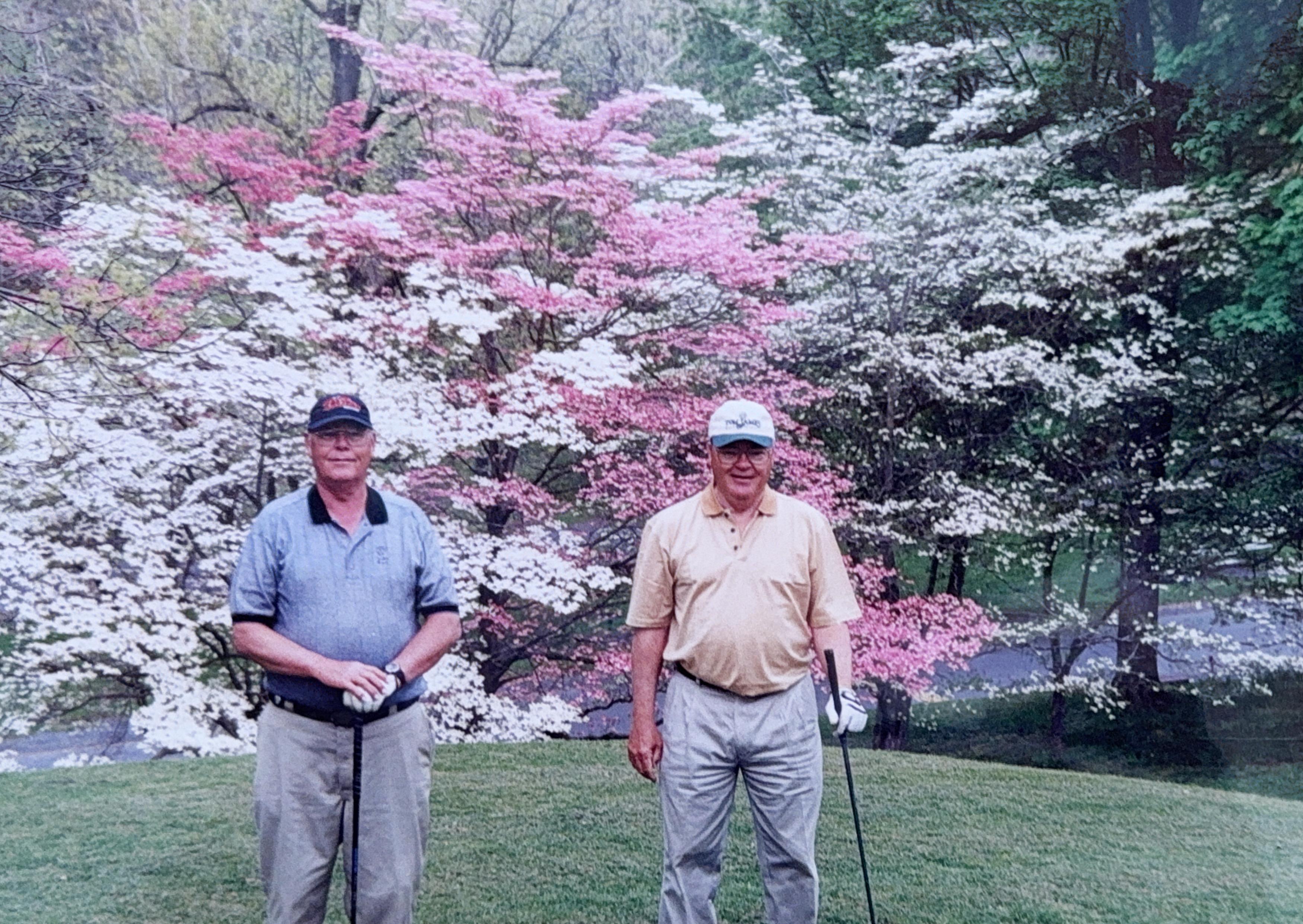 Two friends pose with golf clubs in a scenic location filled with blooming trees on a clear day.