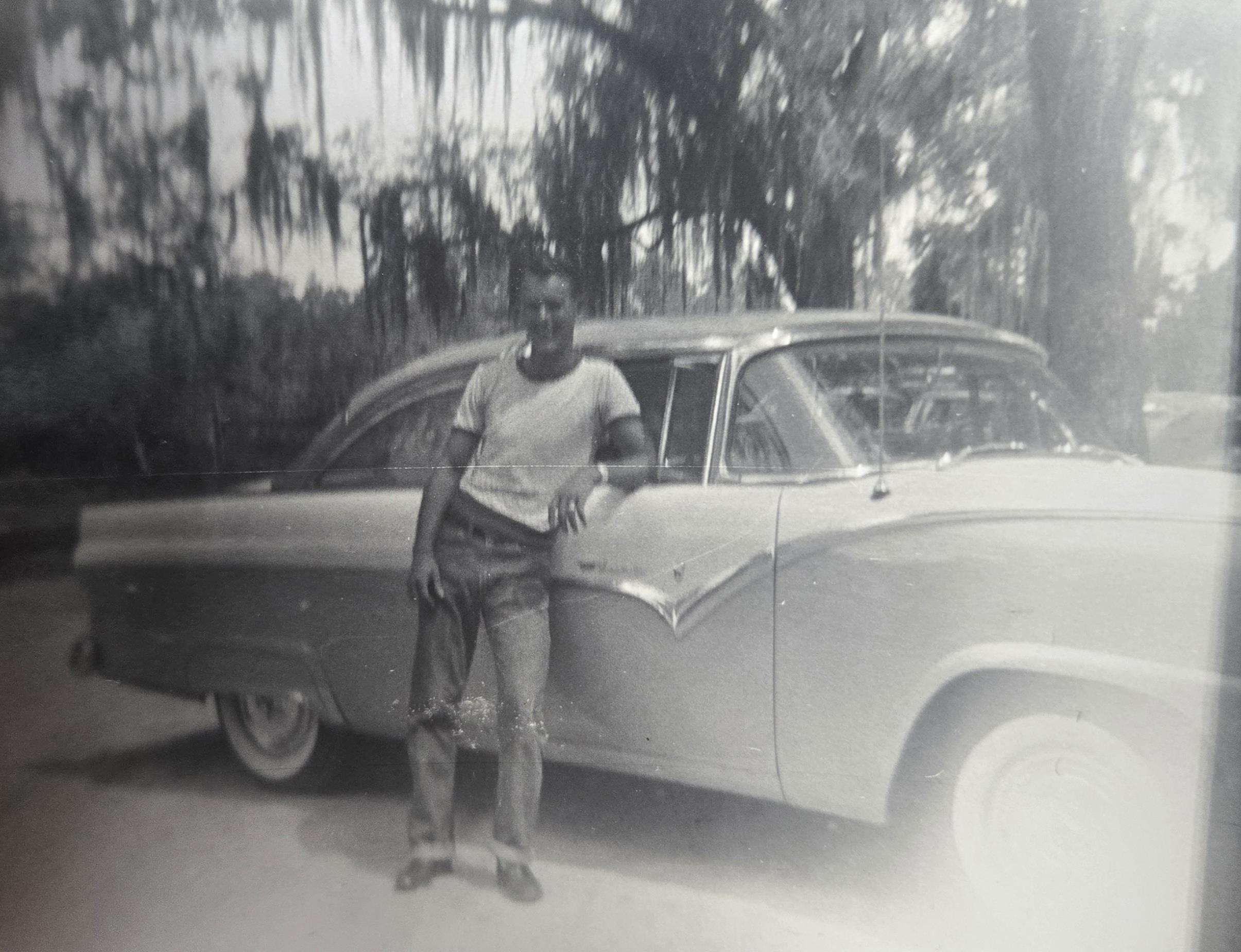 A man stands beside a classic car, showcasing a relaxed attitude in a natural environment.