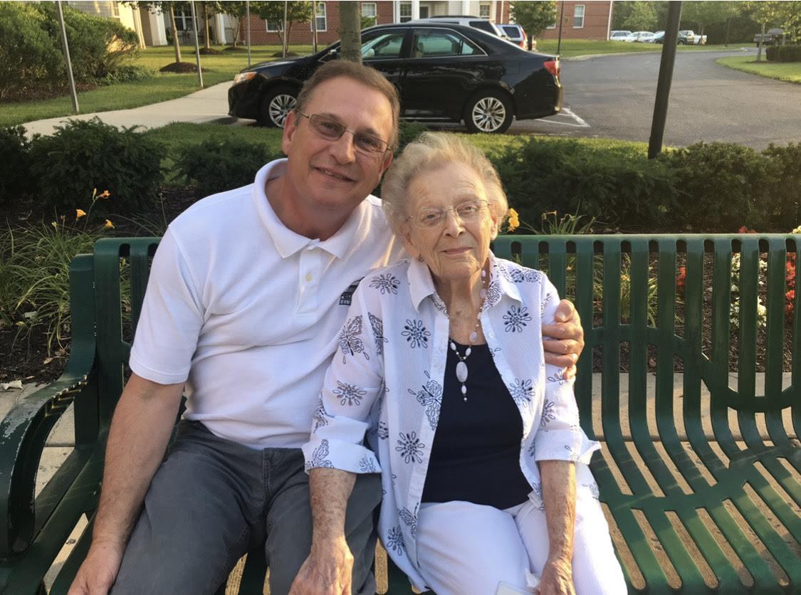 A man sits beside an elderly woman on a green bench, both smiling in the sun.