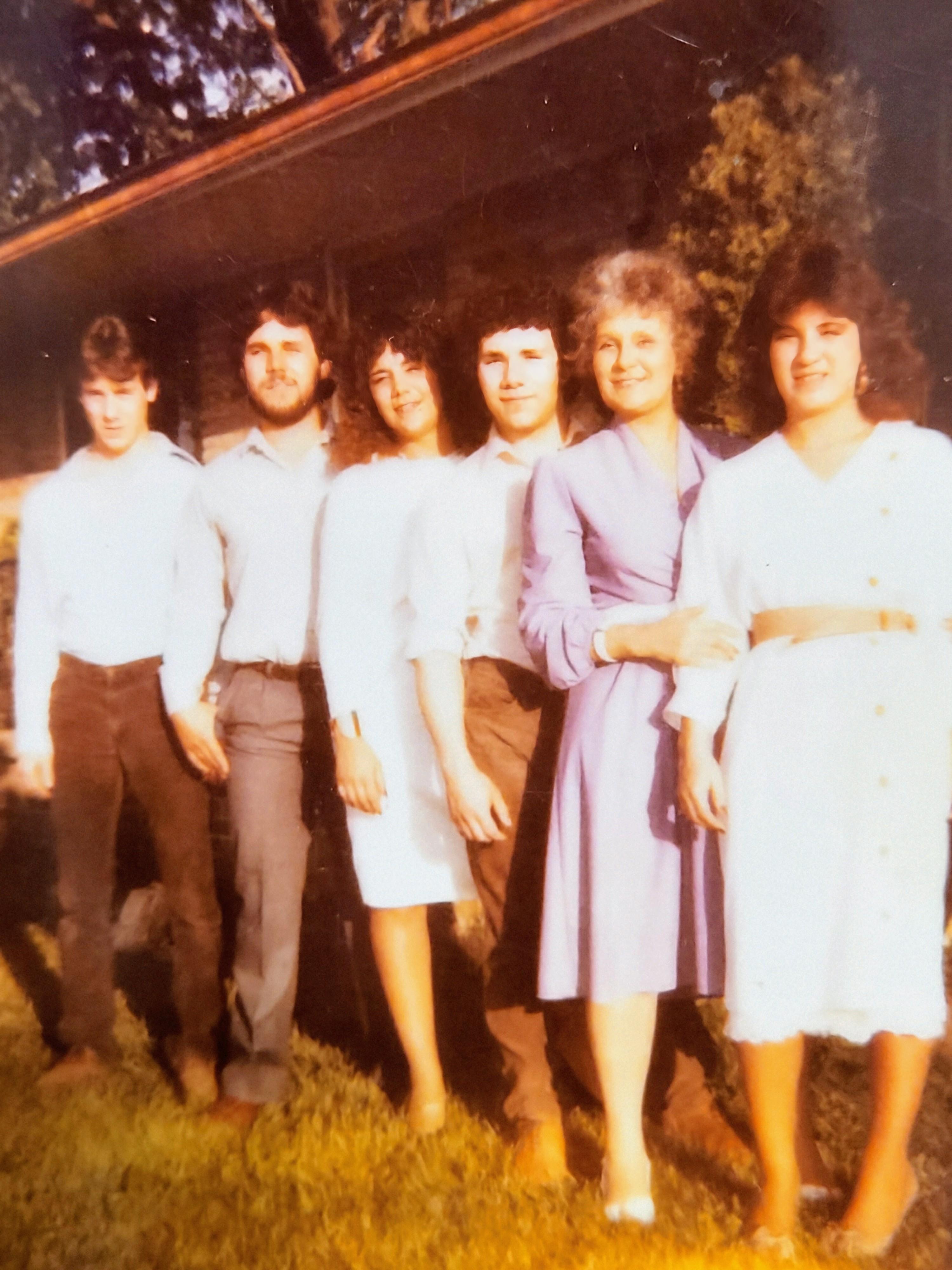 Six friends stand closely together, smiling outdoors by a charming house in bright sunlight.