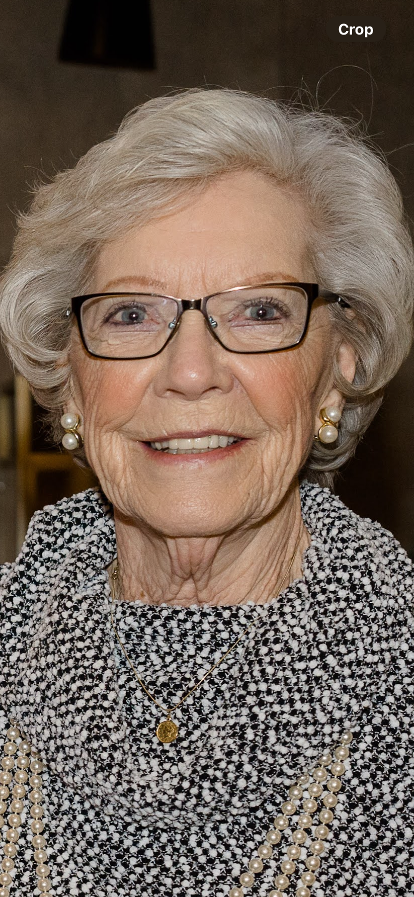 An elderly woman with glasses and pearls smiles warmly at a gathering in a cozy indoor space.