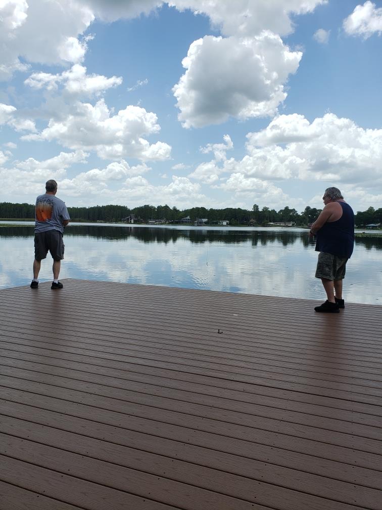 Two individuals stand on a wooden dock beside a serene lake, enjoying a peaceful afternoon.