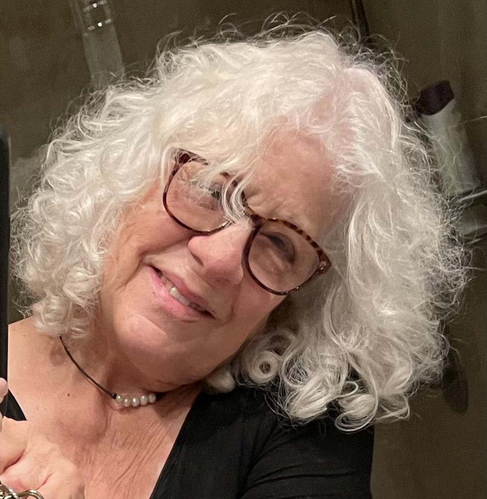 An elderly woman with glasses happily poses with a makeup brush and artistic tools.