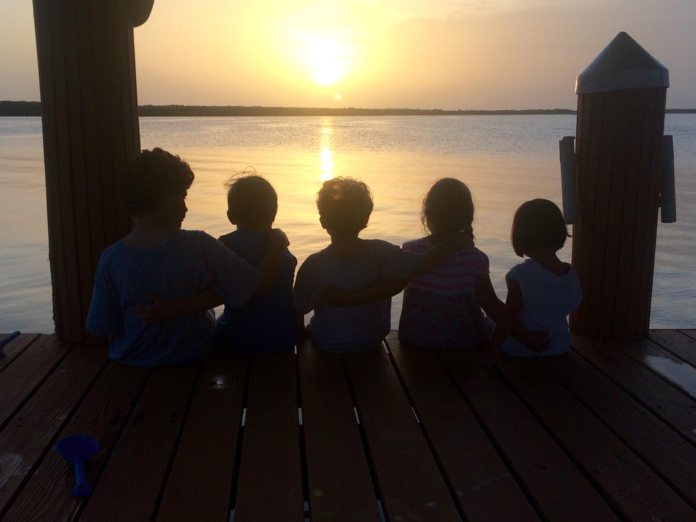 Children sit together on a dock, enjoying a beautiful sunset over the water.