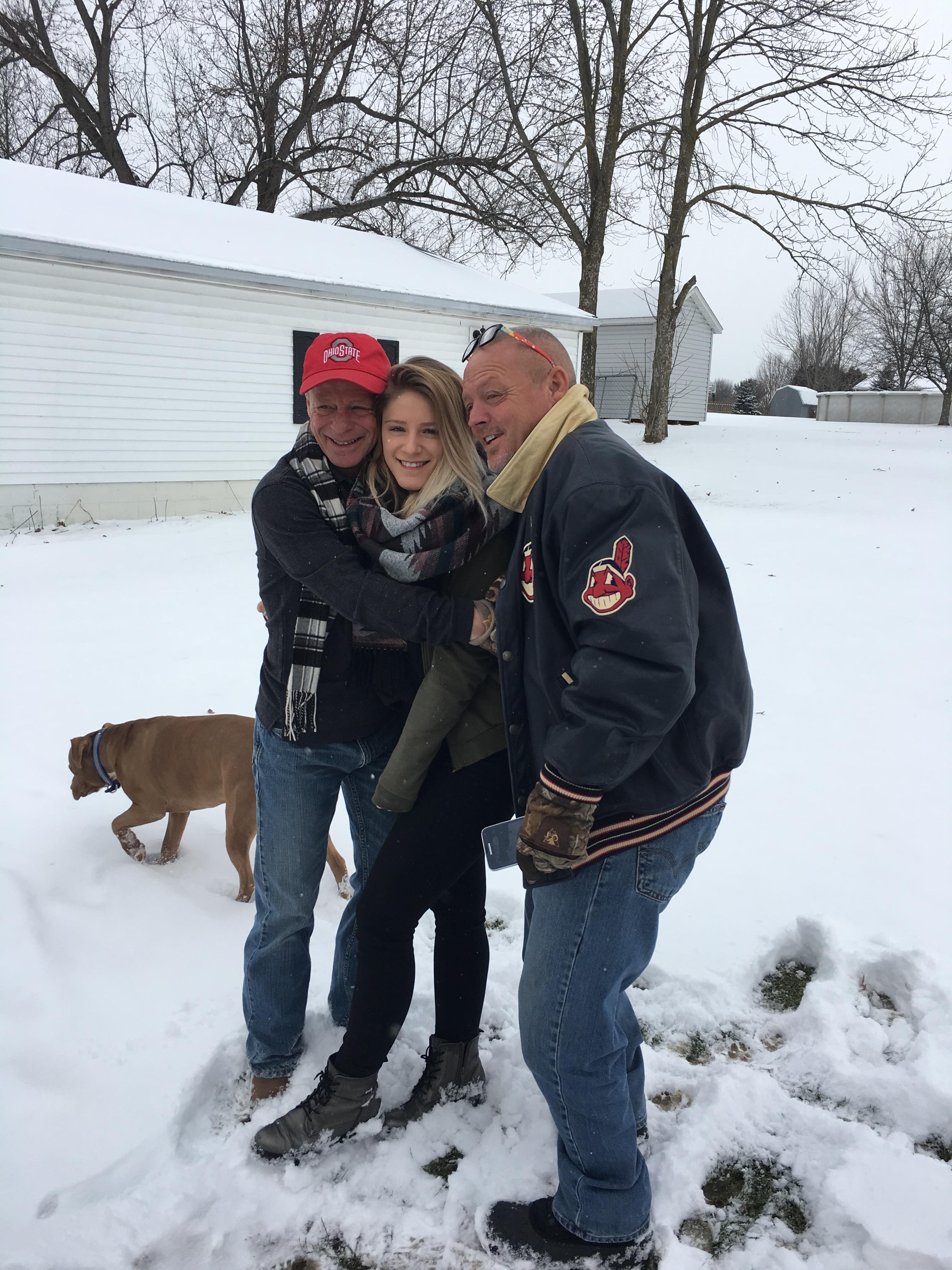 Three people gather in the snow, embracing each other warmly while a dog walks nearby.