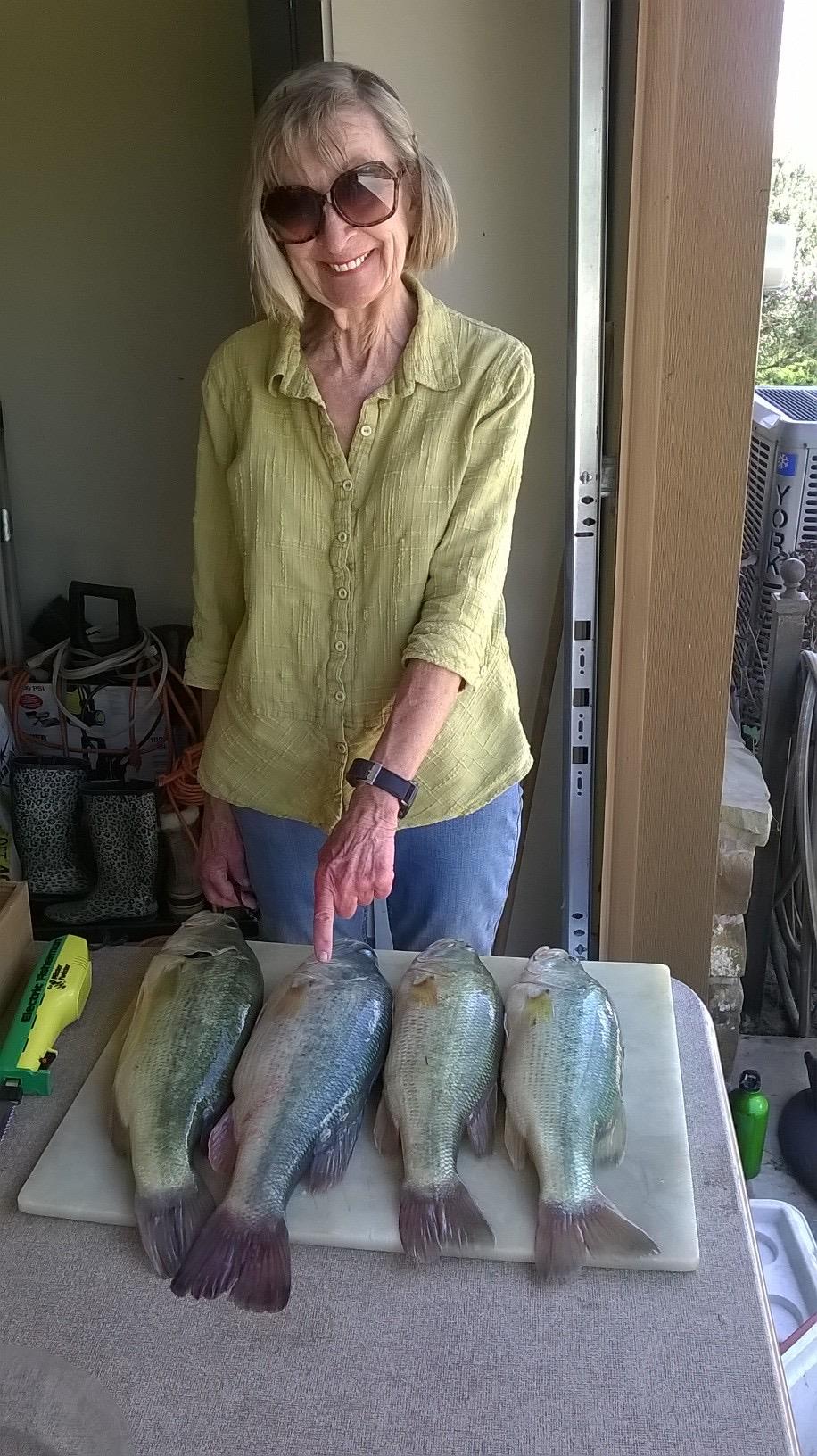 An elderly woman points at several freshly caught fish on a table during a sunny day.