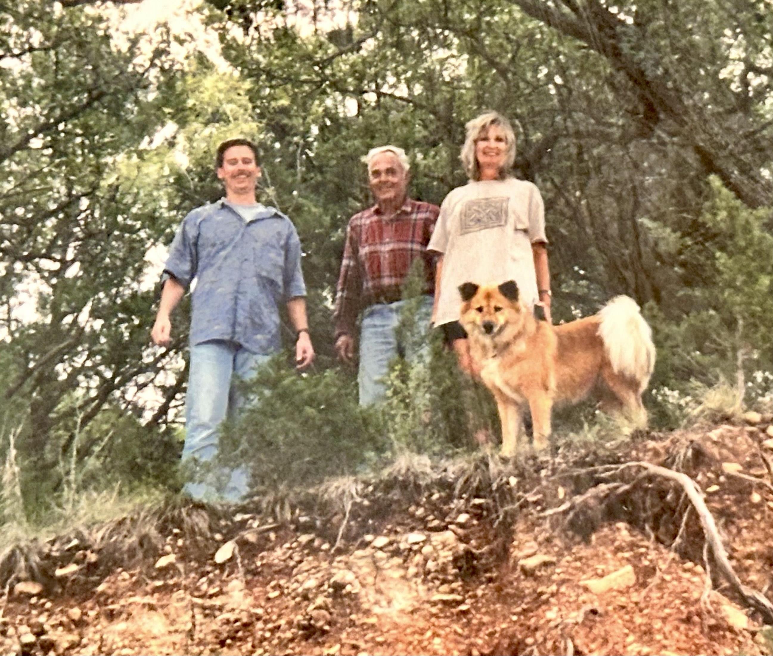 Three individuals pose with a dog on a rocky ledge surrounded by trees in bright light.