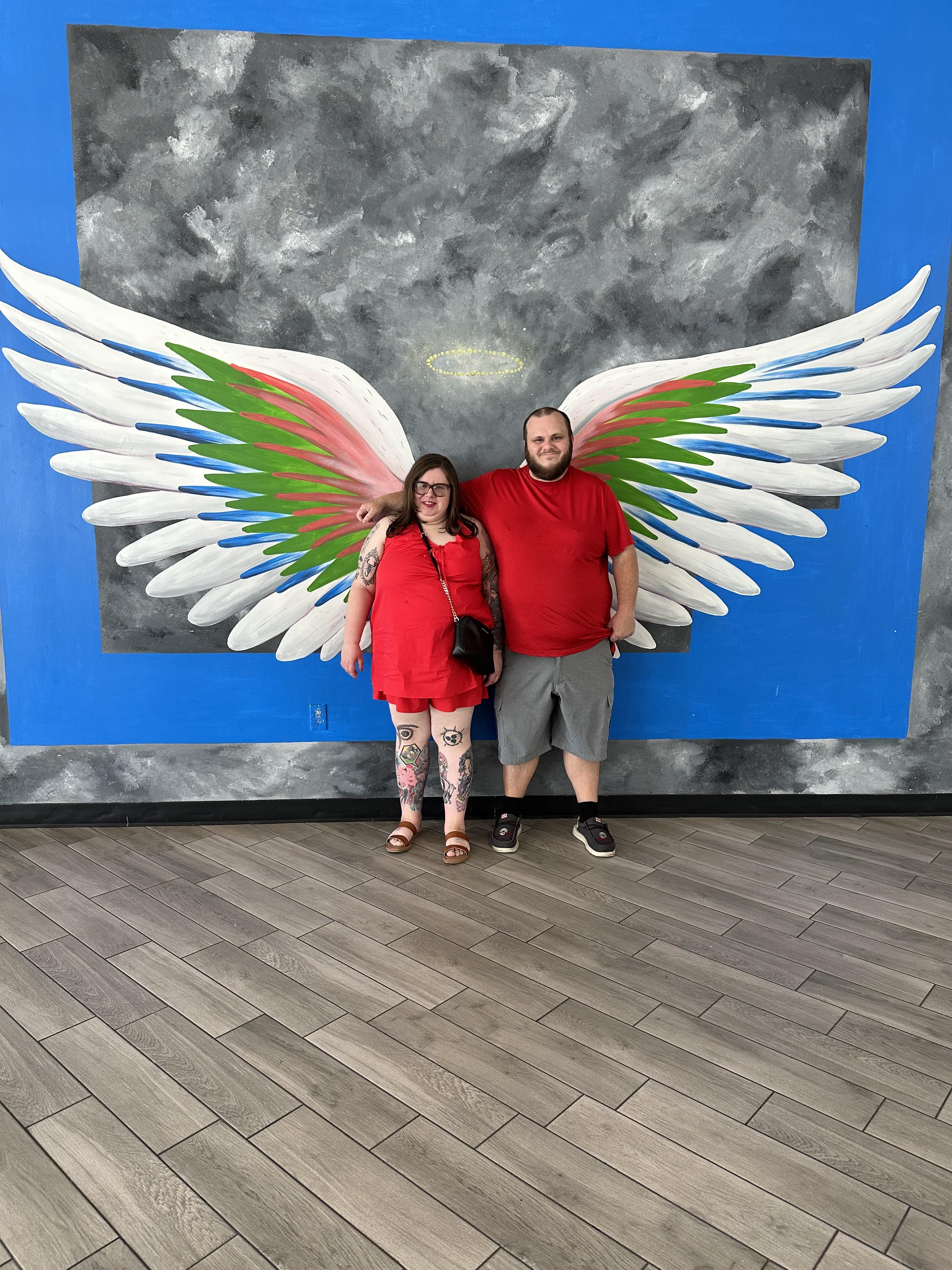 A couple in matching red outfits stands by a colorful wing mural at an art display.