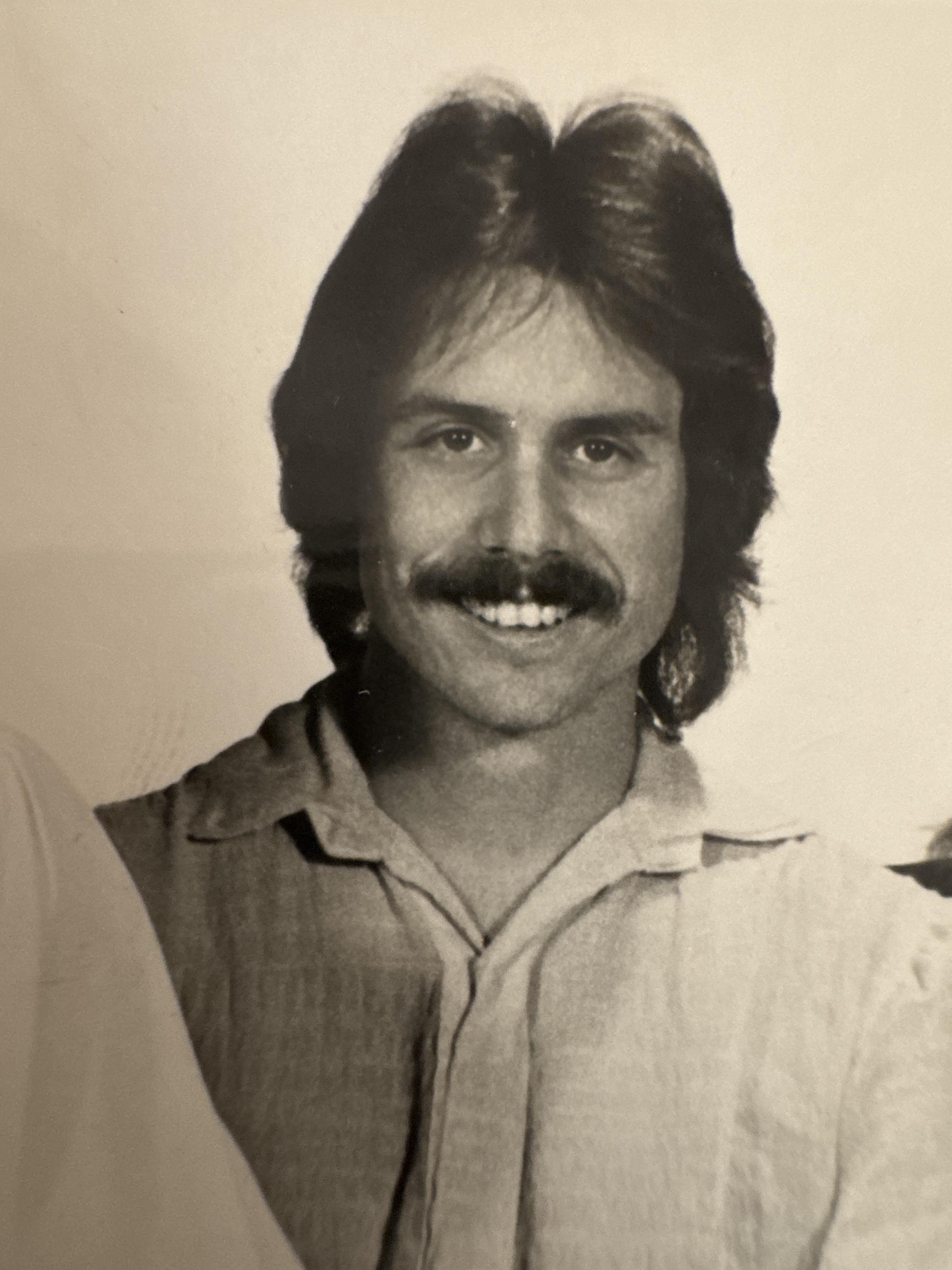 A man with a mustache poses confidently, showcasing a friendly smile and relaxed demeanor.