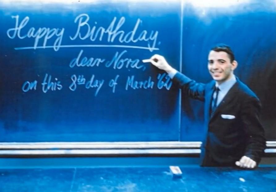 A man in a suit writes a birthday greeting on a blue chalkboard. The date is March 8.