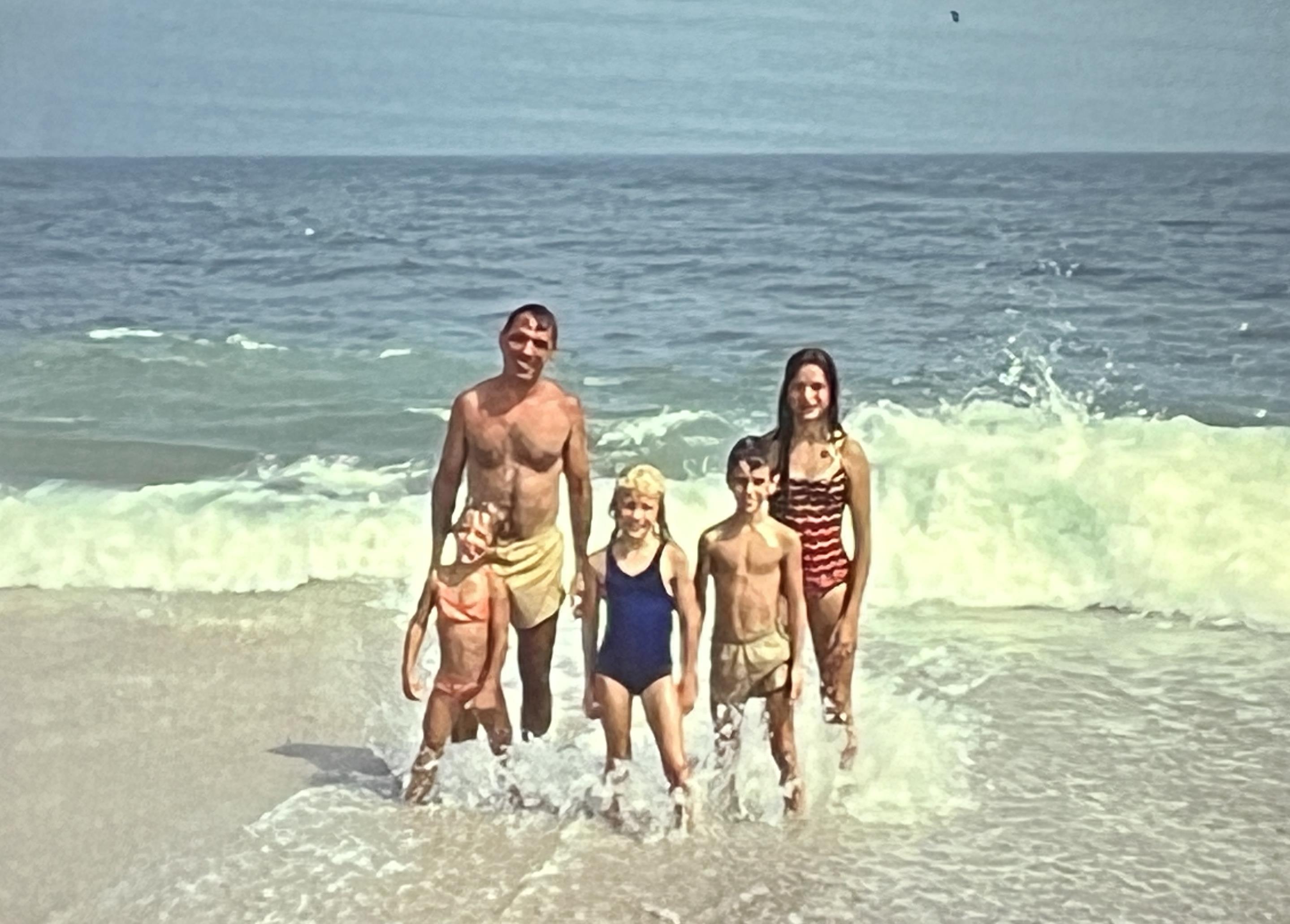 Five family members stand together in the shallow water, smiling and enjoying the beach atmosphere.