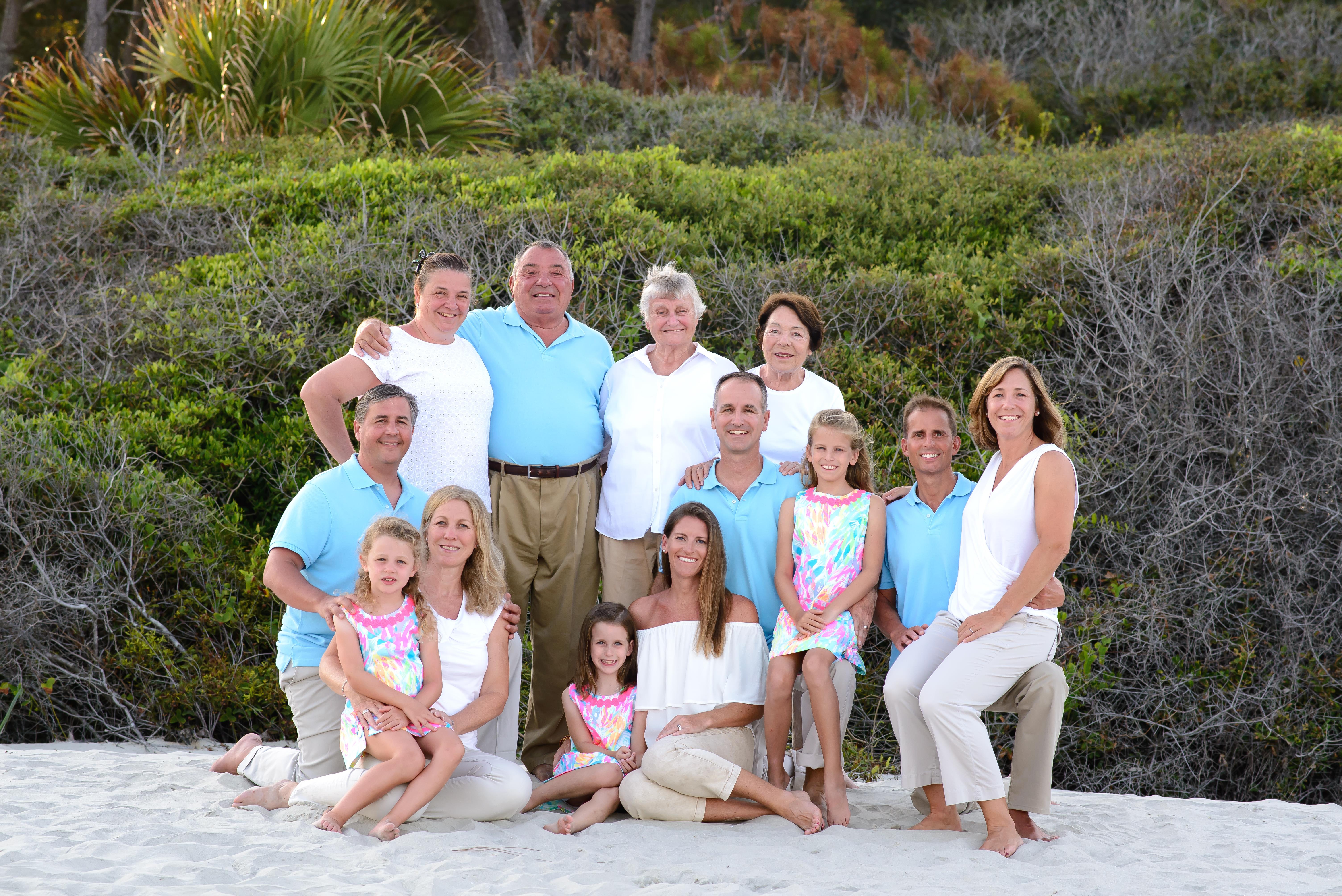 A large family poses together on the beach, smiling and enjoying a sunny day surrounded by greenery.