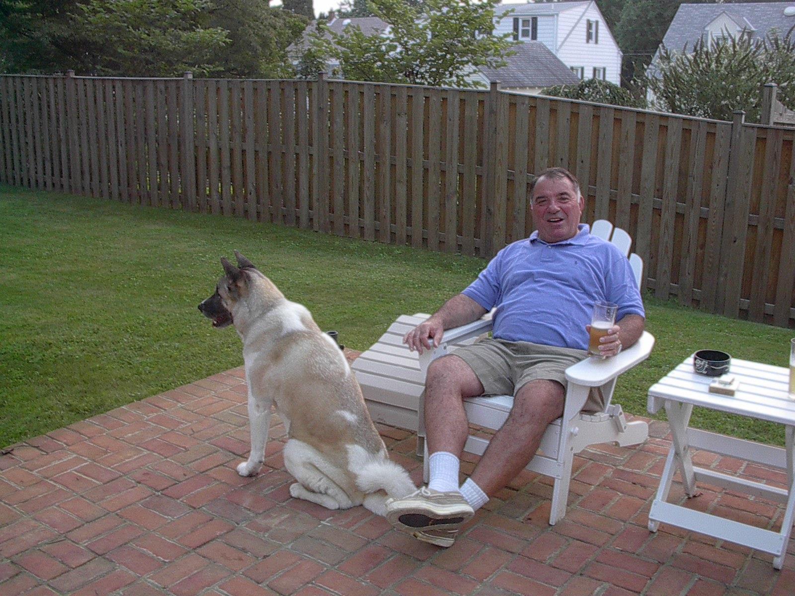 A man sits comfortably in a chair, holding a drink, while his dog rests nearby in a backyard.