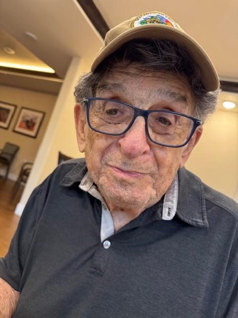 An elderly man wearing glasses and a cap smiles in a relaxed indoor environment with friends.