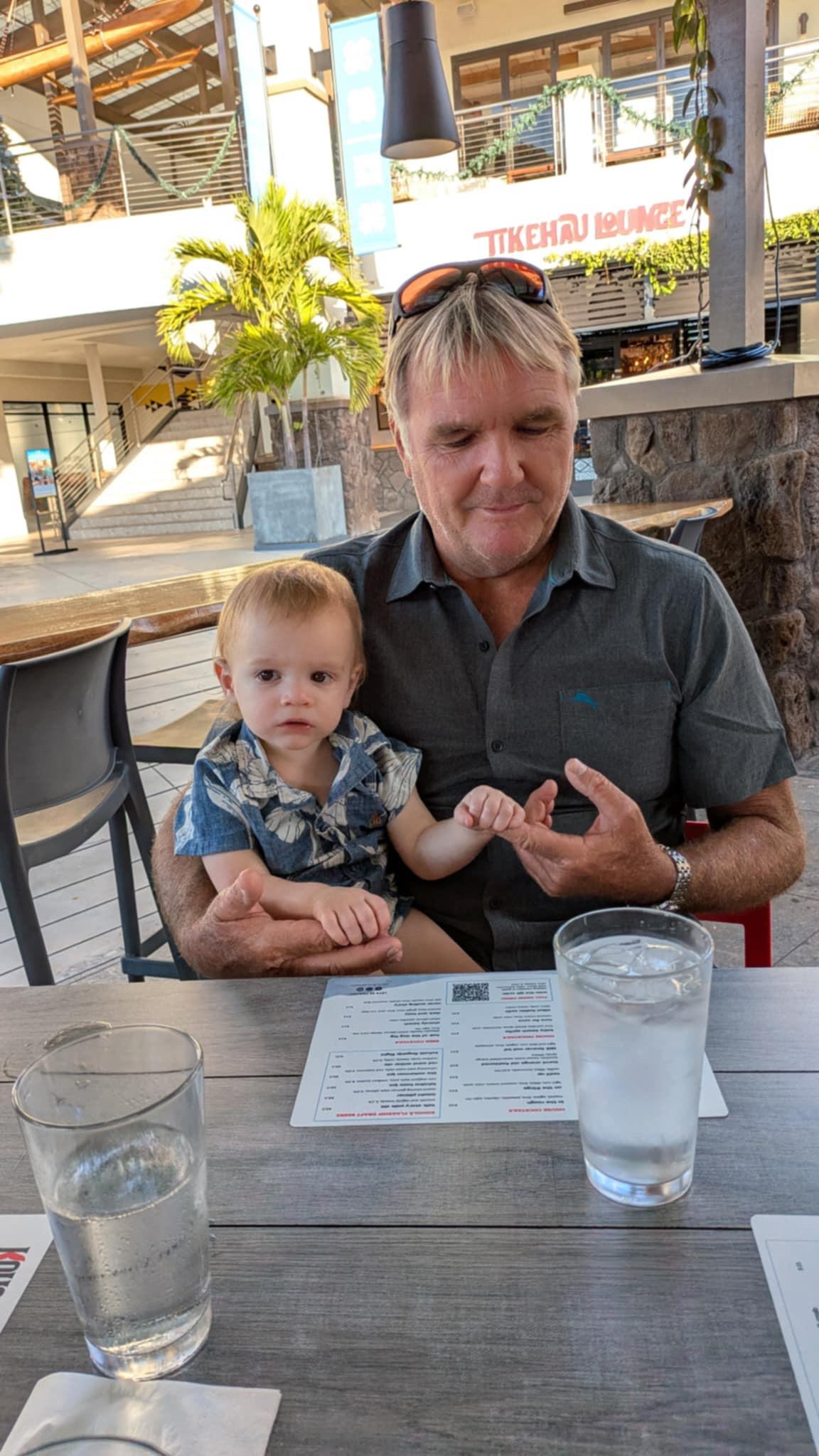 A father enjoys a meal with his young child at an outdoor restaurant, surrounded by nature.
