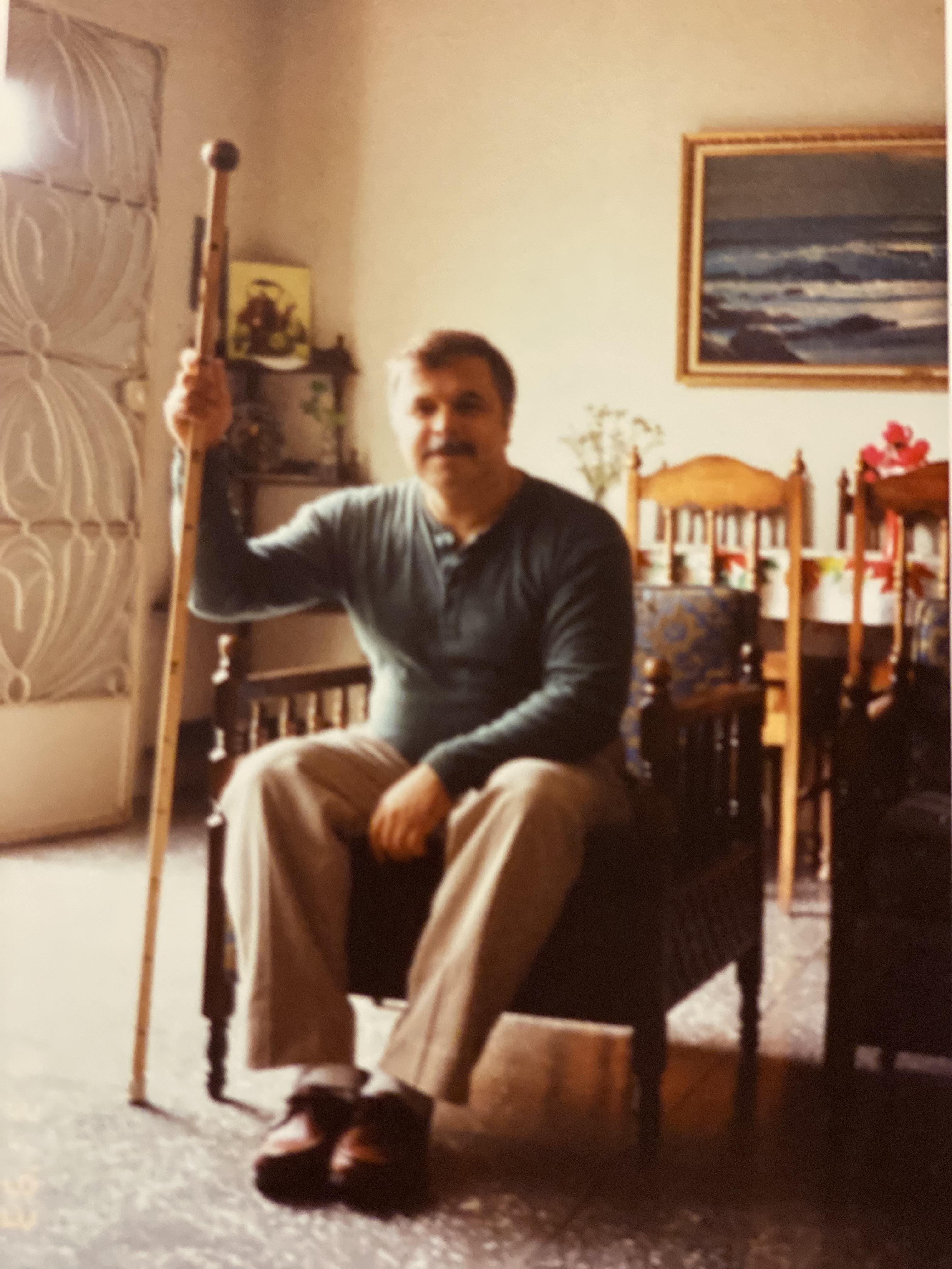 An elderly man sits comfortably in a chair, holding a walking stick while enjoying his surroundings.