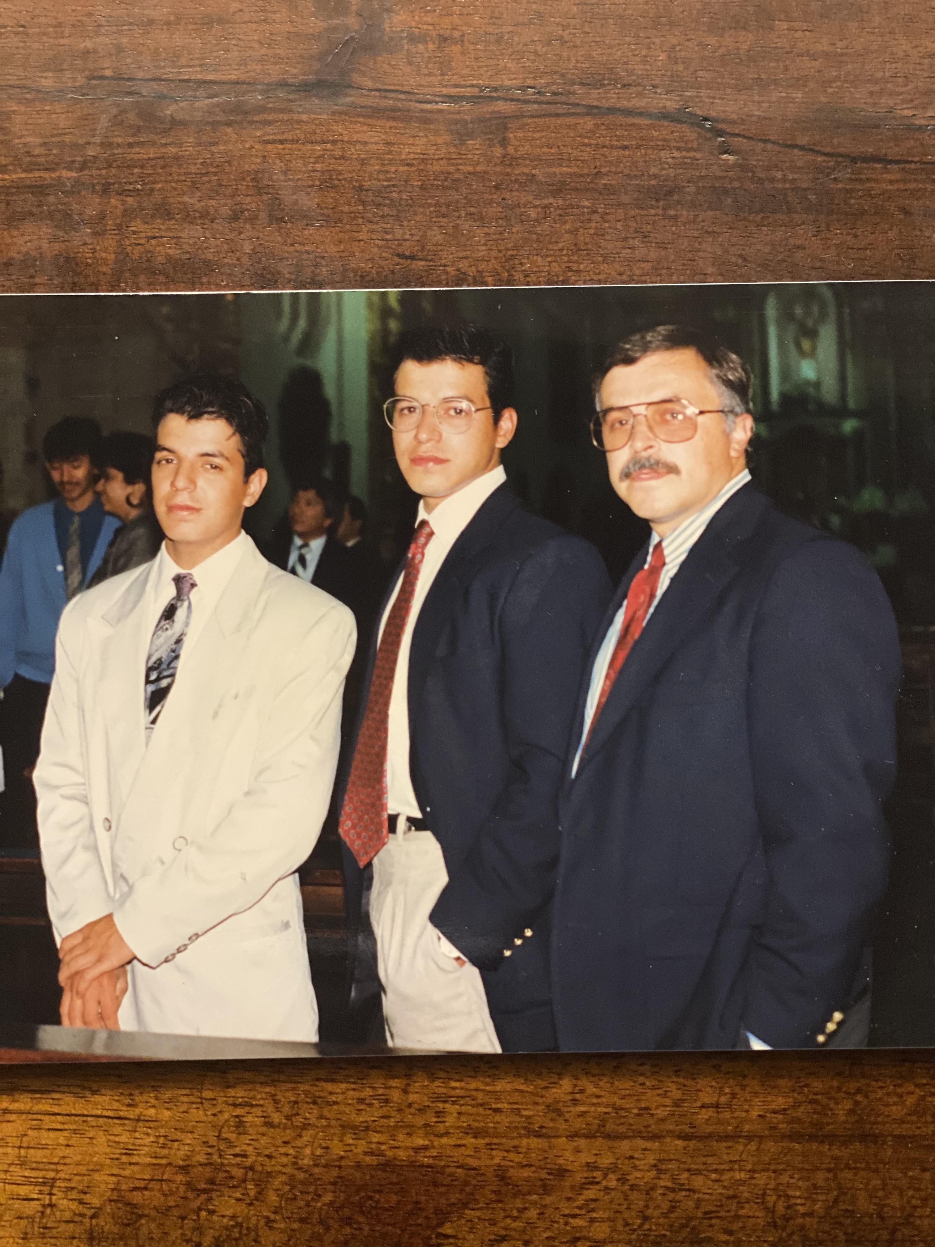 Three men dressed in suits pose confidently at a formal event, surrounded by festive ambiance.